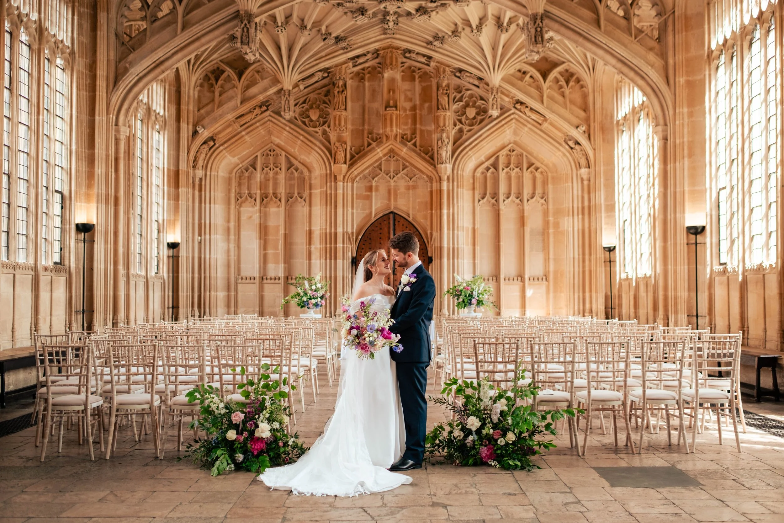A bride and groom standing in the center of an ornate room at their wedding venue, the Bodleian Library in Oxford, surrounded by empty chairs and floral arrangements, sharing a quiet moment on their wedding day.