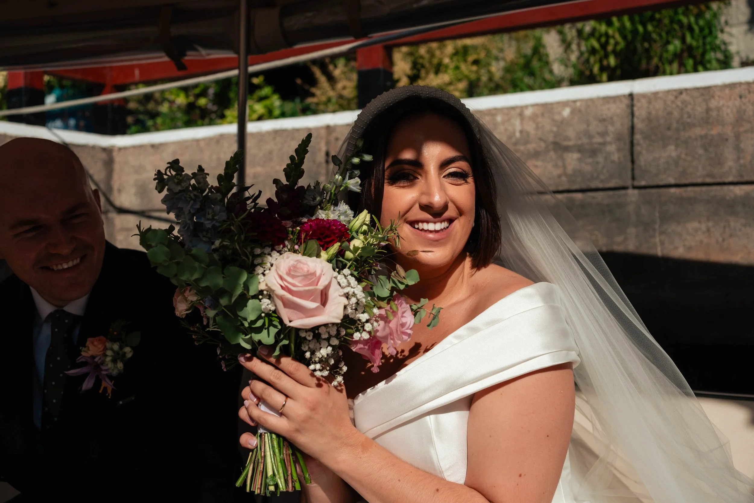 A bride in a white wedding dress and veil holding a bouquet of pink, red, and white flowers, smiling.