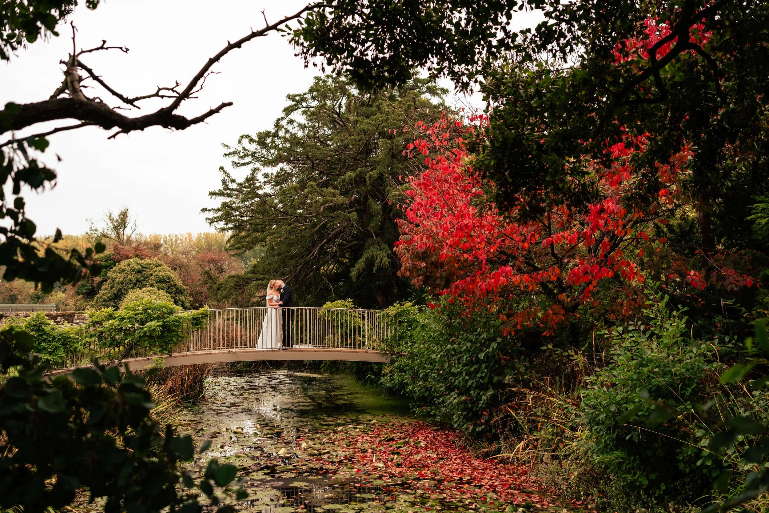 A bride and groom standing together on a small arched bridge over a pond, surrounded by lush trees with green and red leaves during autumn.