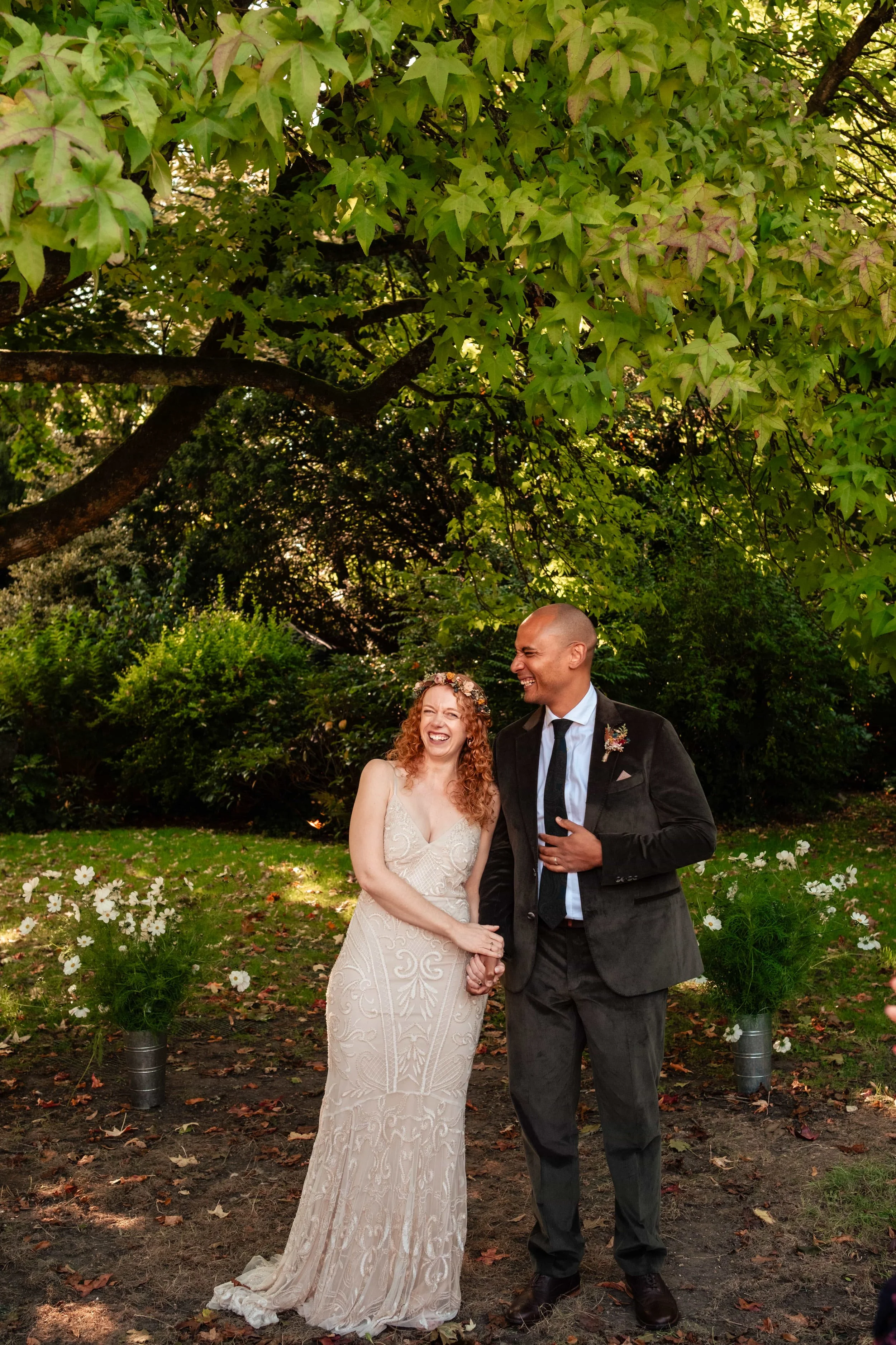 A newlywed couple stands together outdoors under a large leafy tree, smiling and laughing, with a background of greenery and white flowers in vases.