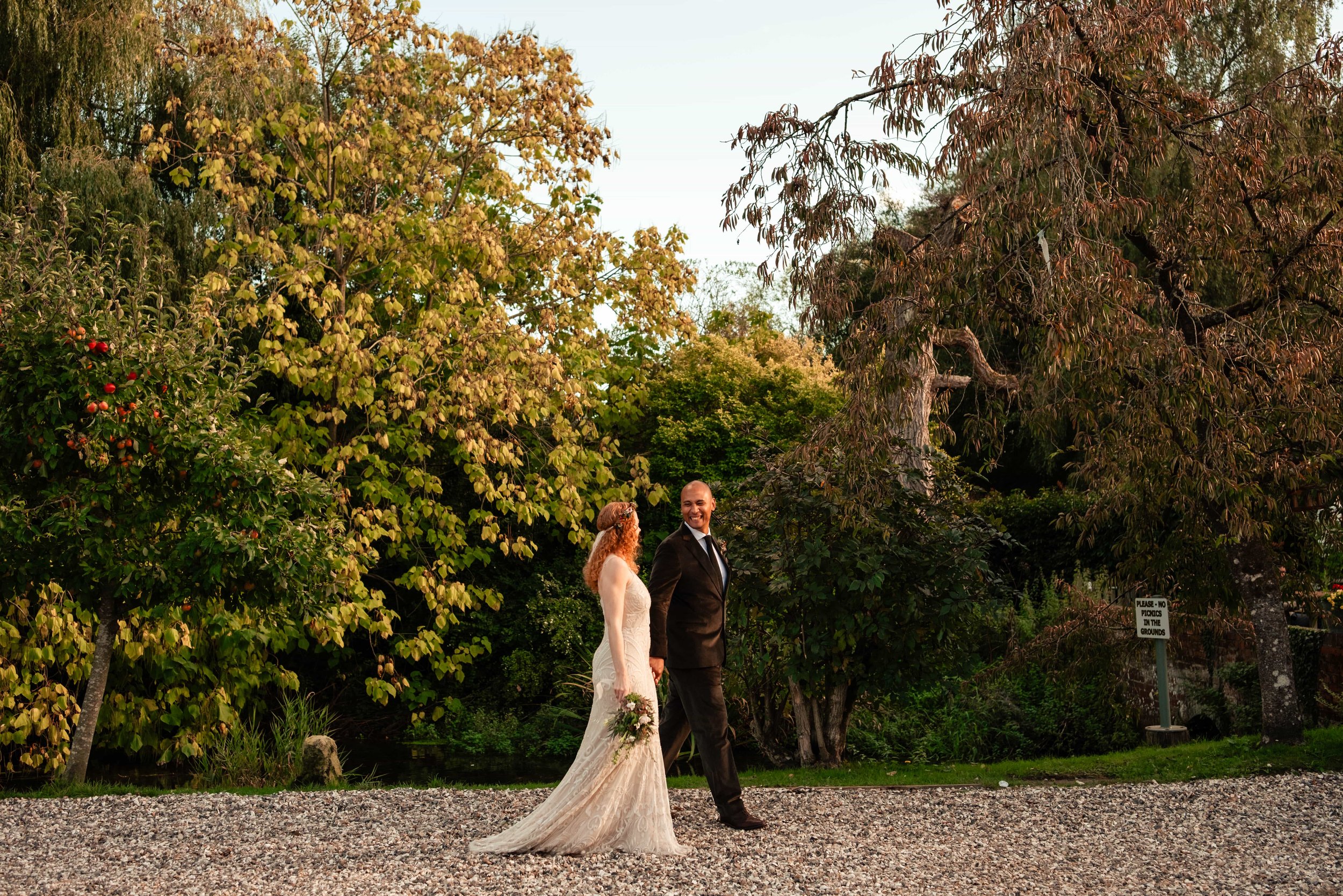 A bride and groom walking outdoors on a gravel path in a park, holding hands. The bride is wearing a white lace wedding dress and holding a bouquet, while the groom is dressed in a black suit with a tie. The background features lush green trees with 