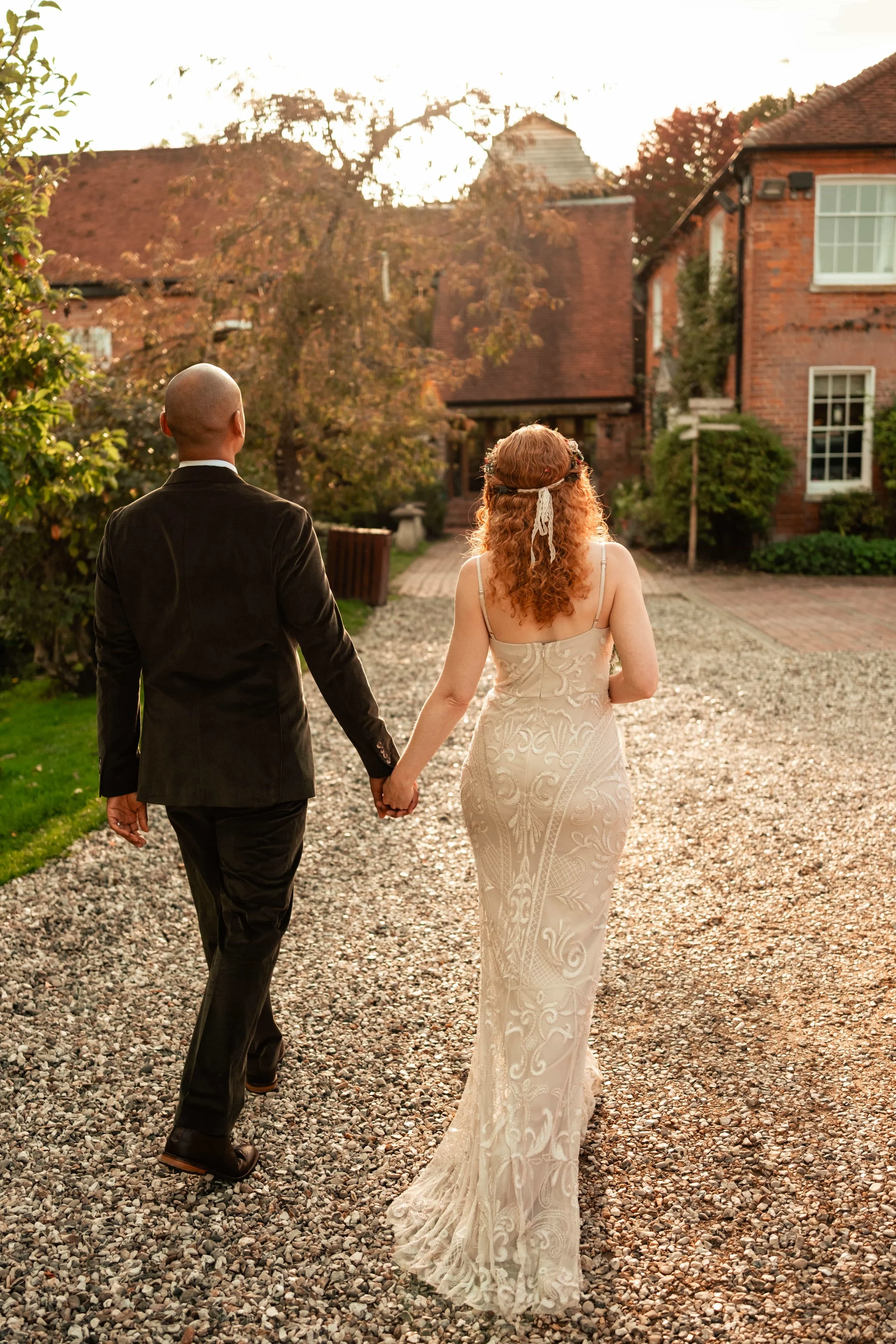 A couple dressed in wedding attire walking hand-in-hand outdoors during sunset in a garden area with brick houses and trees in the background.
