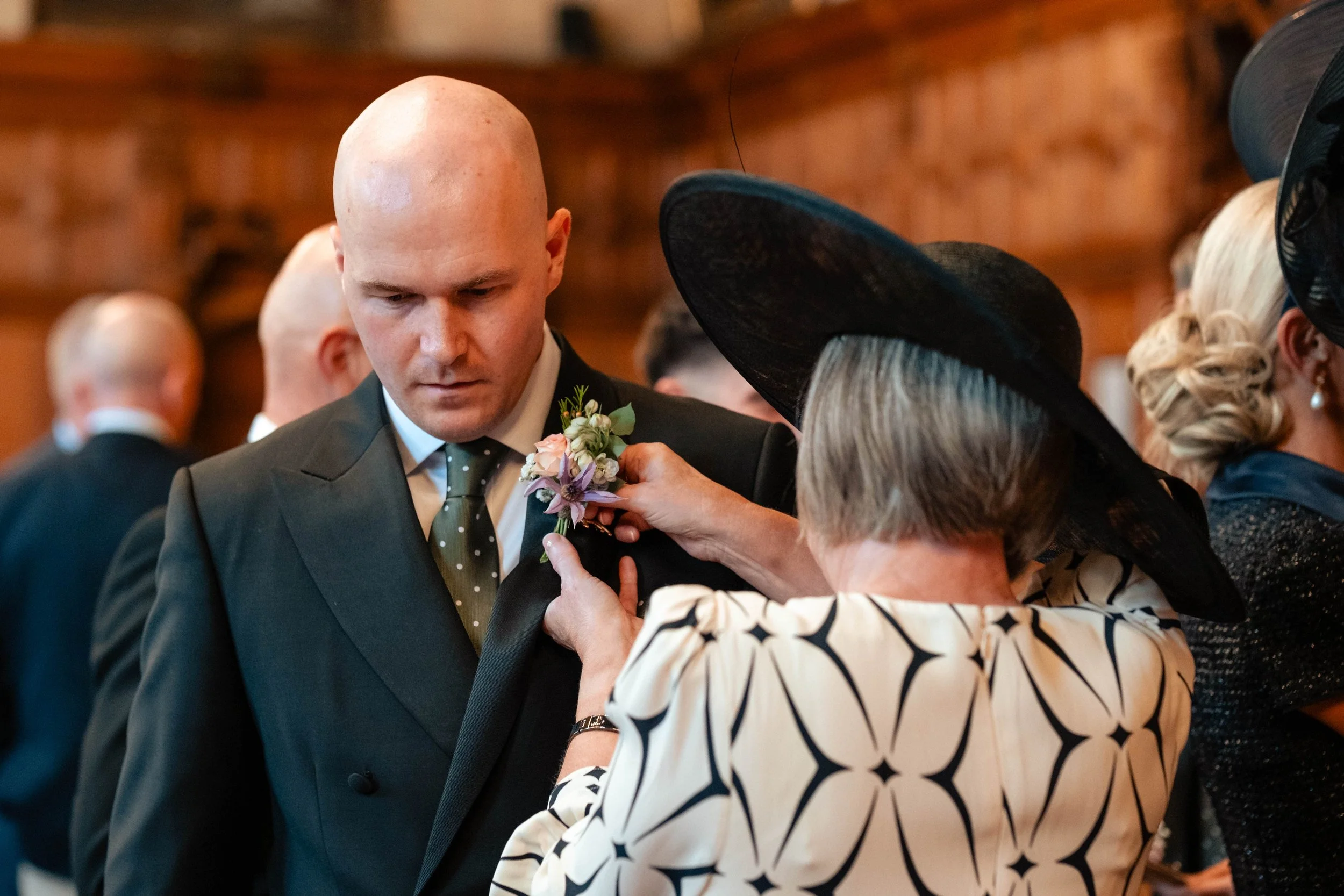A woman in a large black hat pins a purple and white boutonniere onto the lapel of the groom wearing a black suit at a wedding with other people visible in the background in Oxford Town Hall.