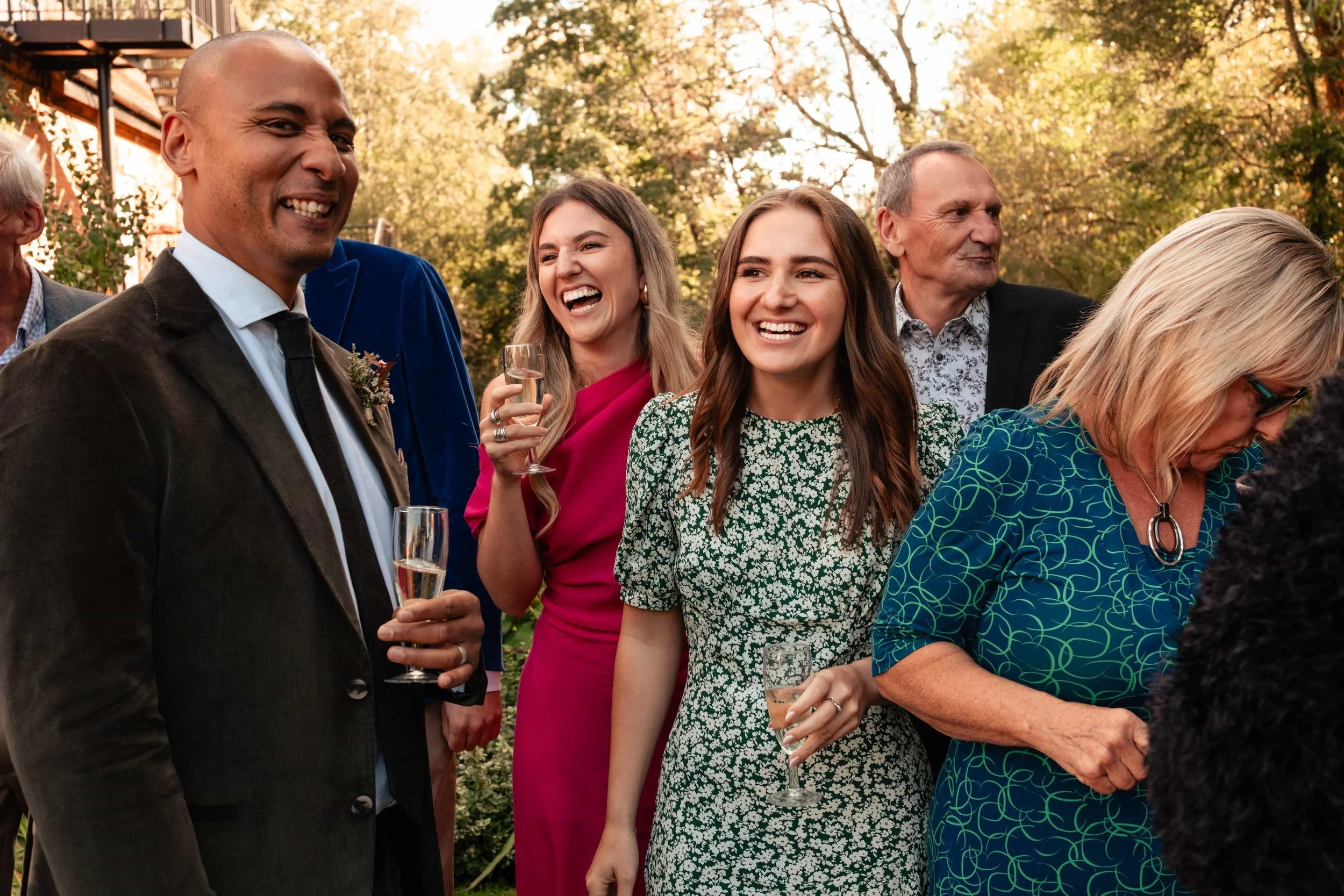 People at a social gathering outdoors, holding glasses of champagne and smiling, with trees and a building in the background.