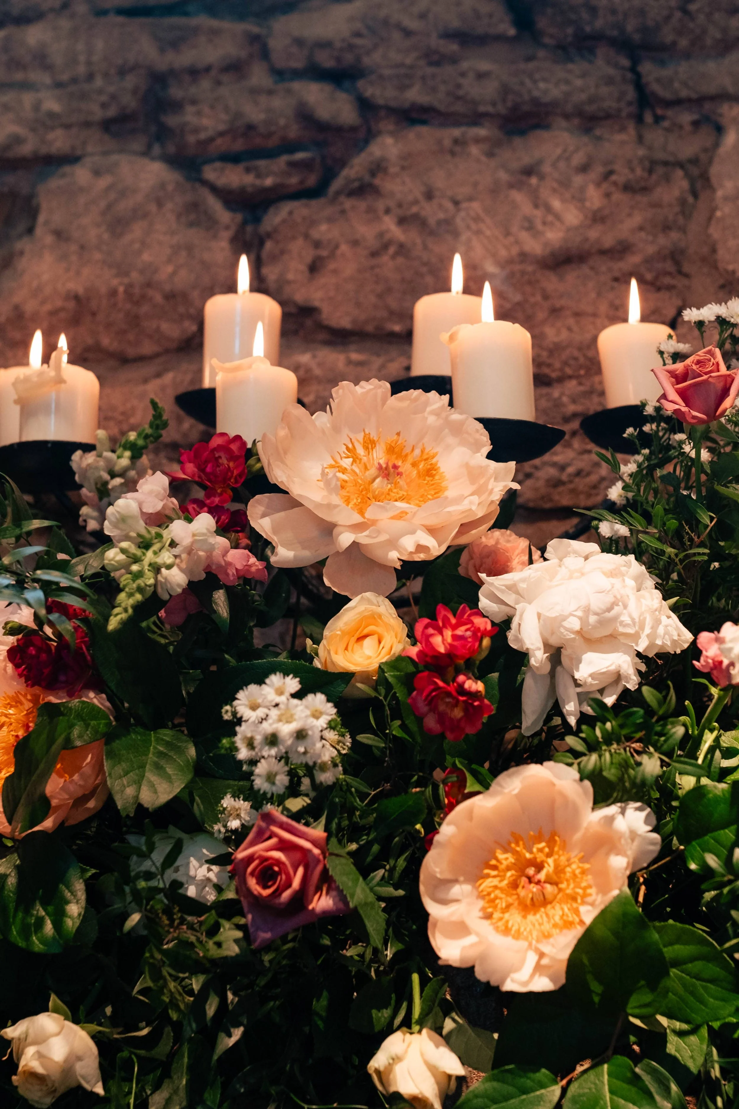 A close-up of a floral arrangement with white, pink, and red flowers, topped with white candles on black holders, against a stone wall background.