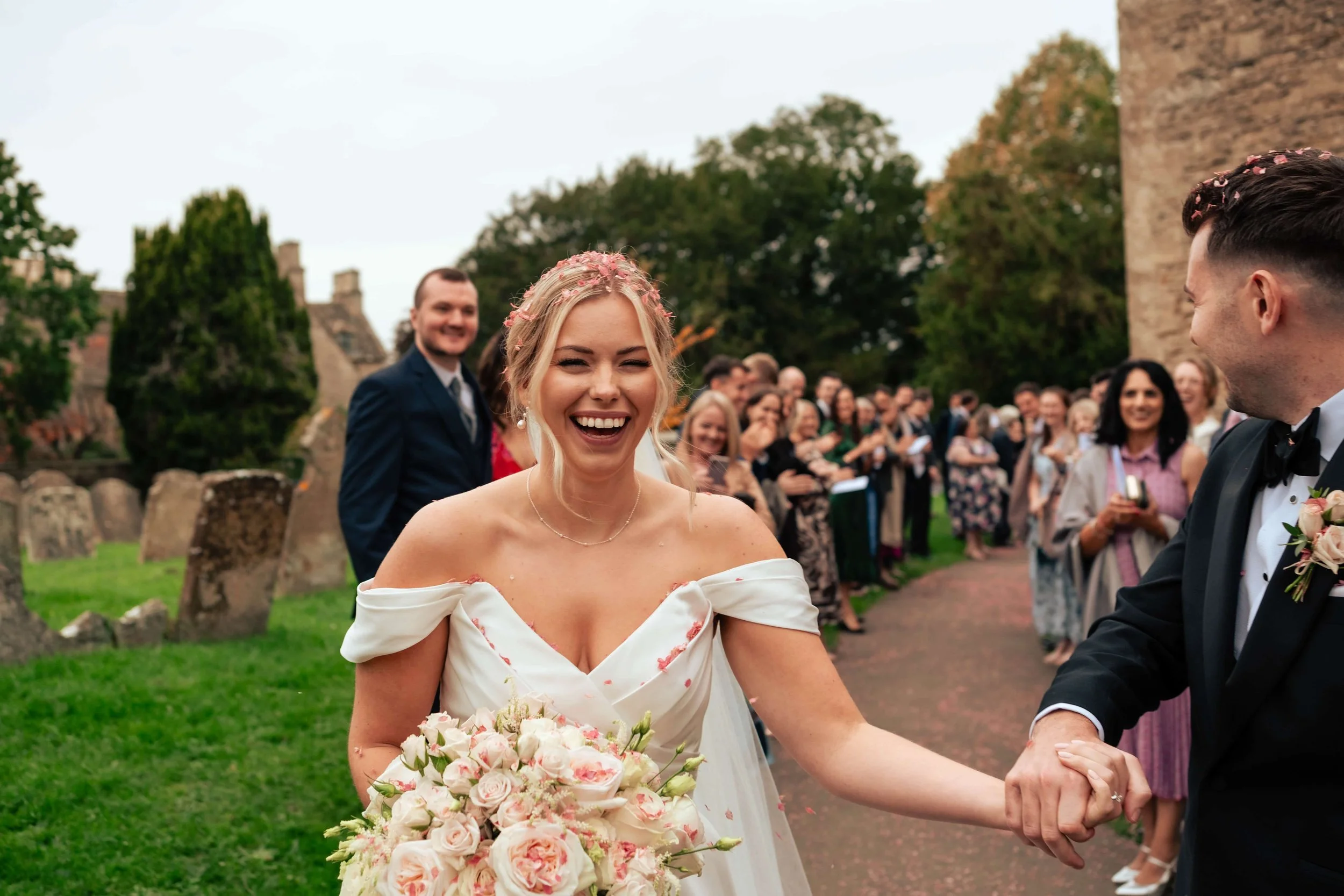 A bride in an off-the-shoulder wedding dress holding a bouquet of pink and white flowers, smiling and holding hands with a groom in a tuxedo during a wedding celebration outdoors, with guests and trees in the background.