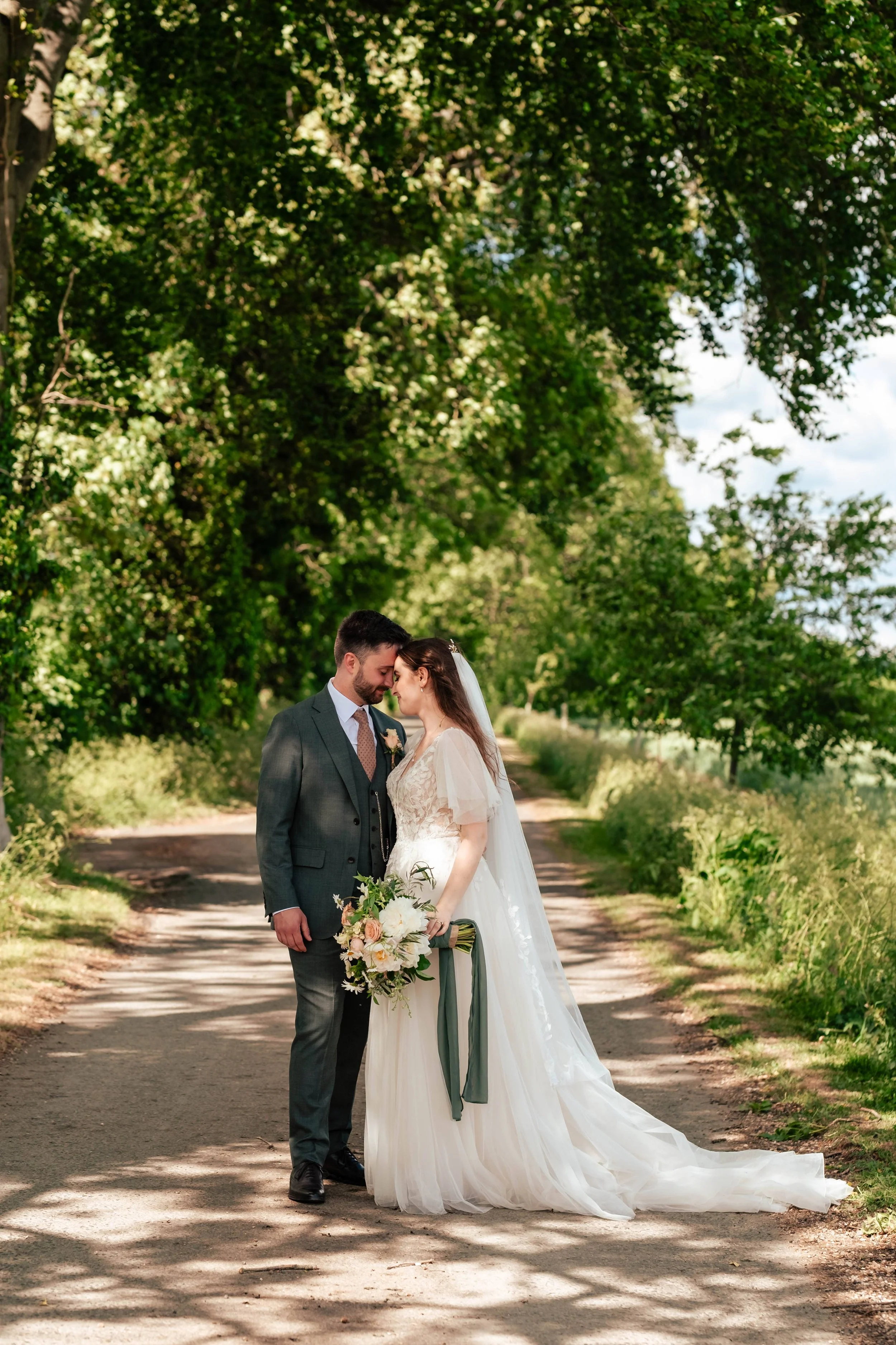 A bride and groom standing close together on a dirt path surrounded by lush green trees, sharing a tender moment, with the bride holding a bouquet of flowers.