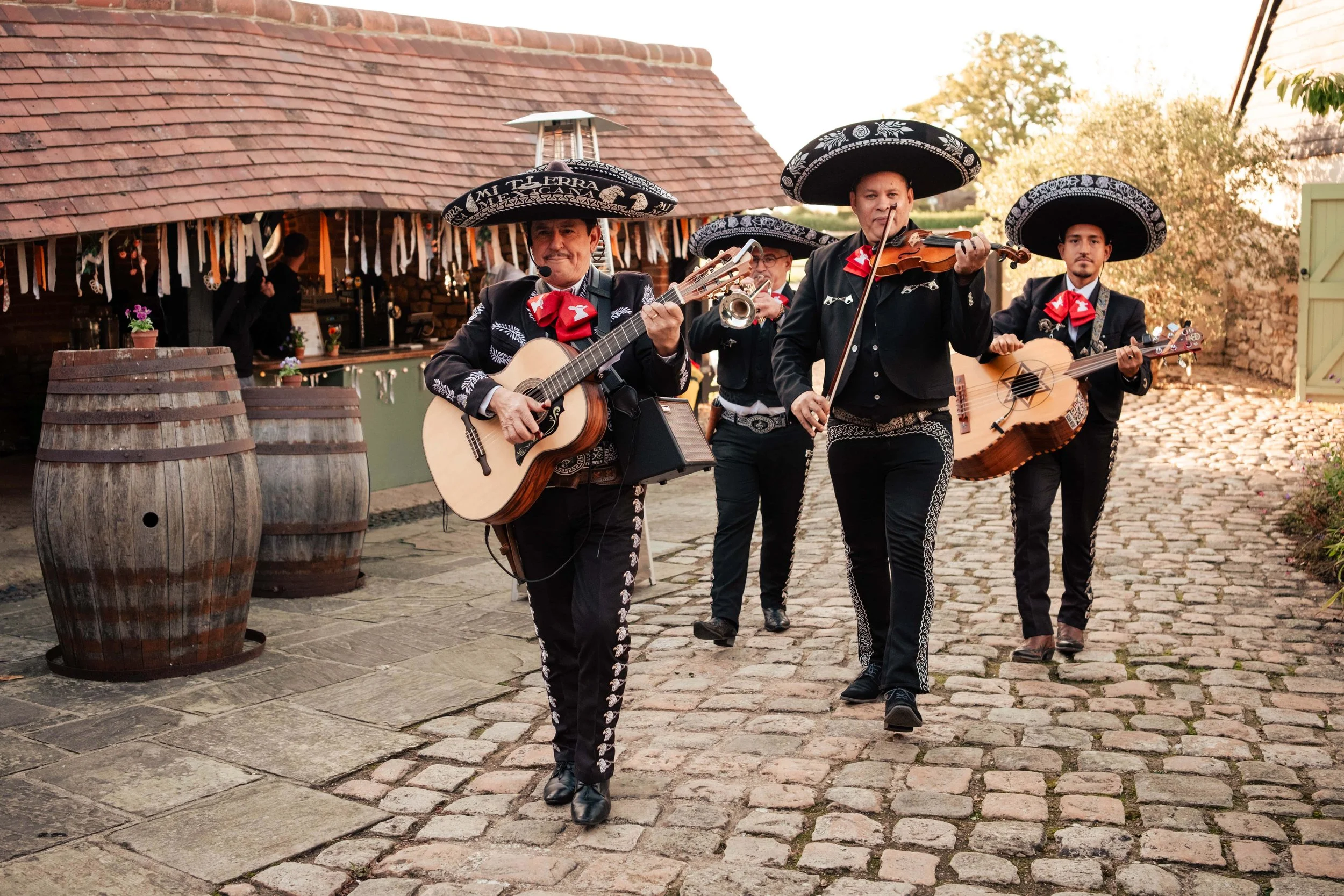 Mariachi band performing outdoors on cobblestone path, wearing traditional black mariachi outfits with sombreros, playing guitars, violin, and trumpet.