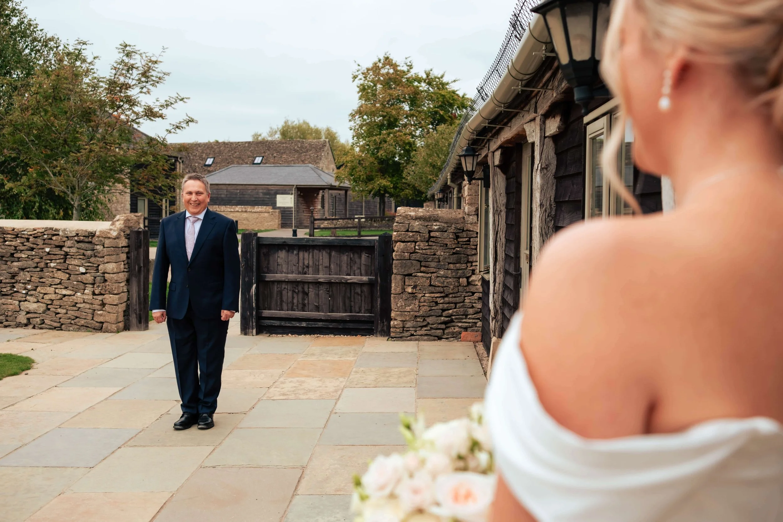 The father of the bride in a suit walking towards a bride outdoors on a patio, holding a bouquet of roses, with a stone wall and rustic building in the background.