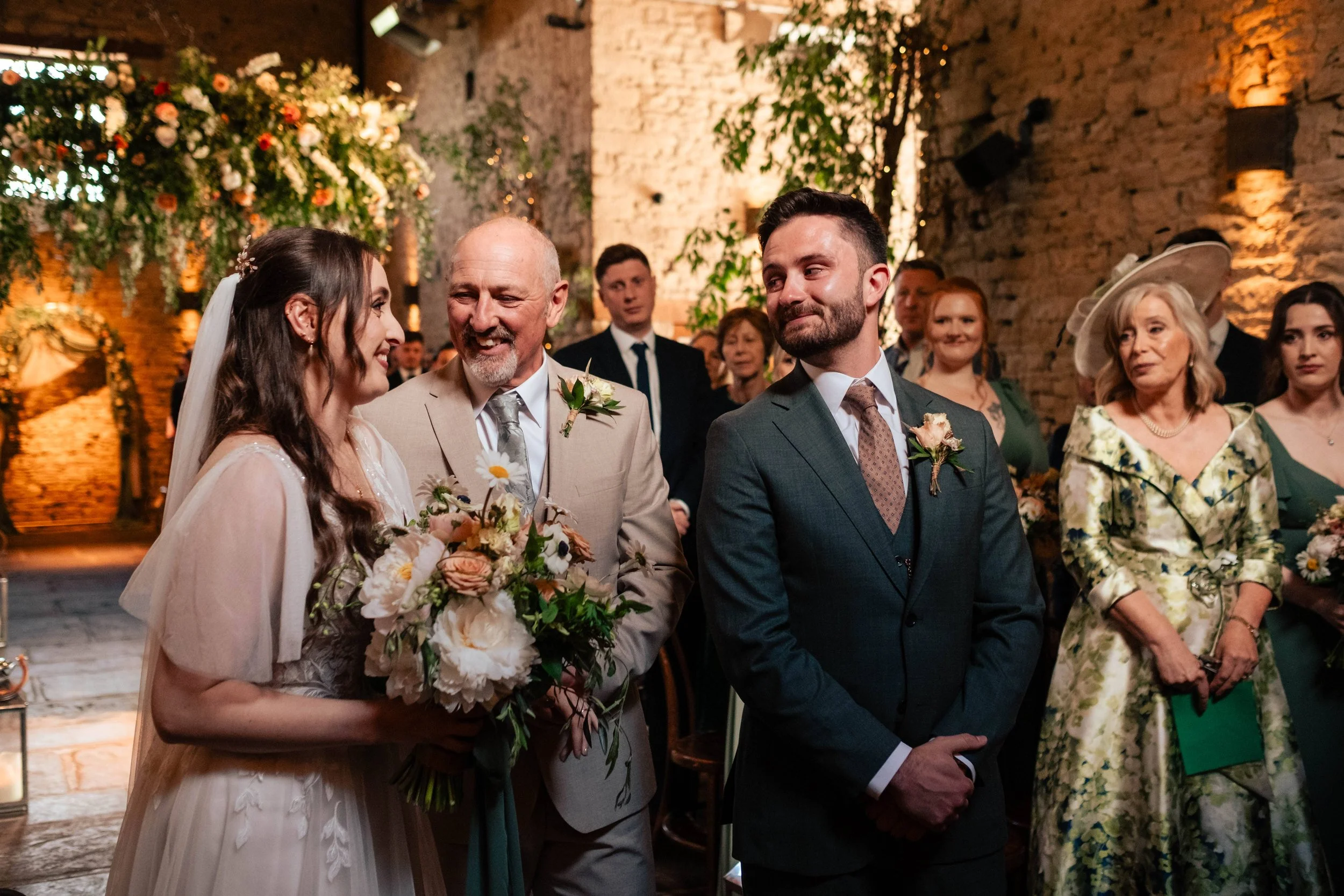 A wedding ceremony with a bride holding a bouquet of flowers, standing next to an older man, possibly her father, and a groom in a dark suit. Guests are gathered in a rustic venue with exposed brick walls and warm lighting, watching the ceremony.