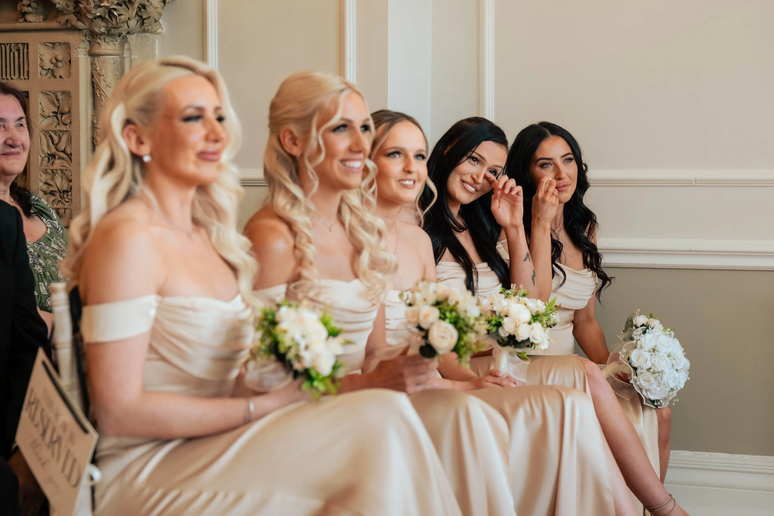 Bridesmaid wiping away tears as she sits with the bridal party in pink-gold dresses along the aisle at Eastington Park wedding venue.