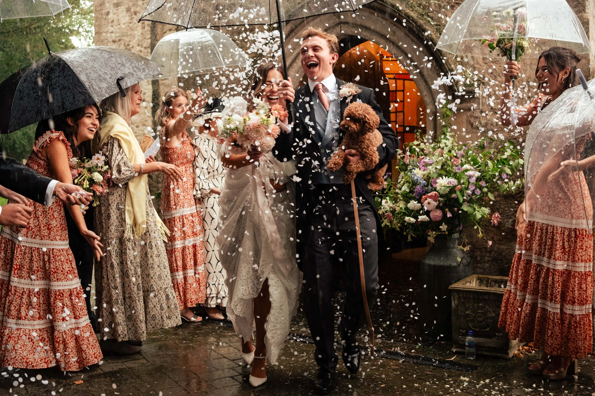 A wedding celebration with a bride and groom walking arm in arm amidst a shower of confetti, surrounded by friends and family holding umbrellas and flowers outside a stone building.