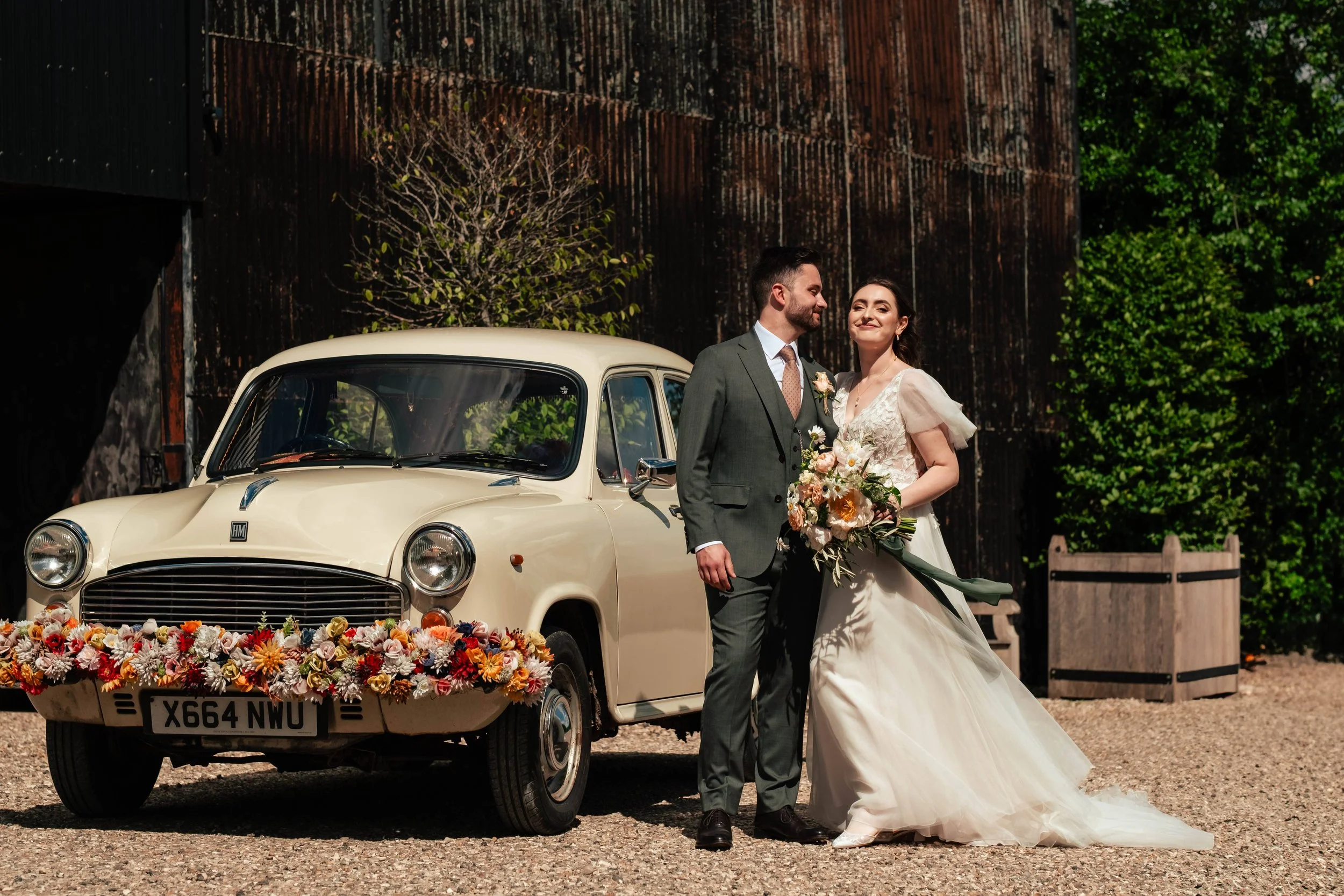 A bride and groom standing next to a vintage cream-colored car decorated with a colorful floral garland on the front. The bride is holding a bouquet and smiling, and the groom is looking at her. They are outdoors with greenery and a rustic wooden wal
