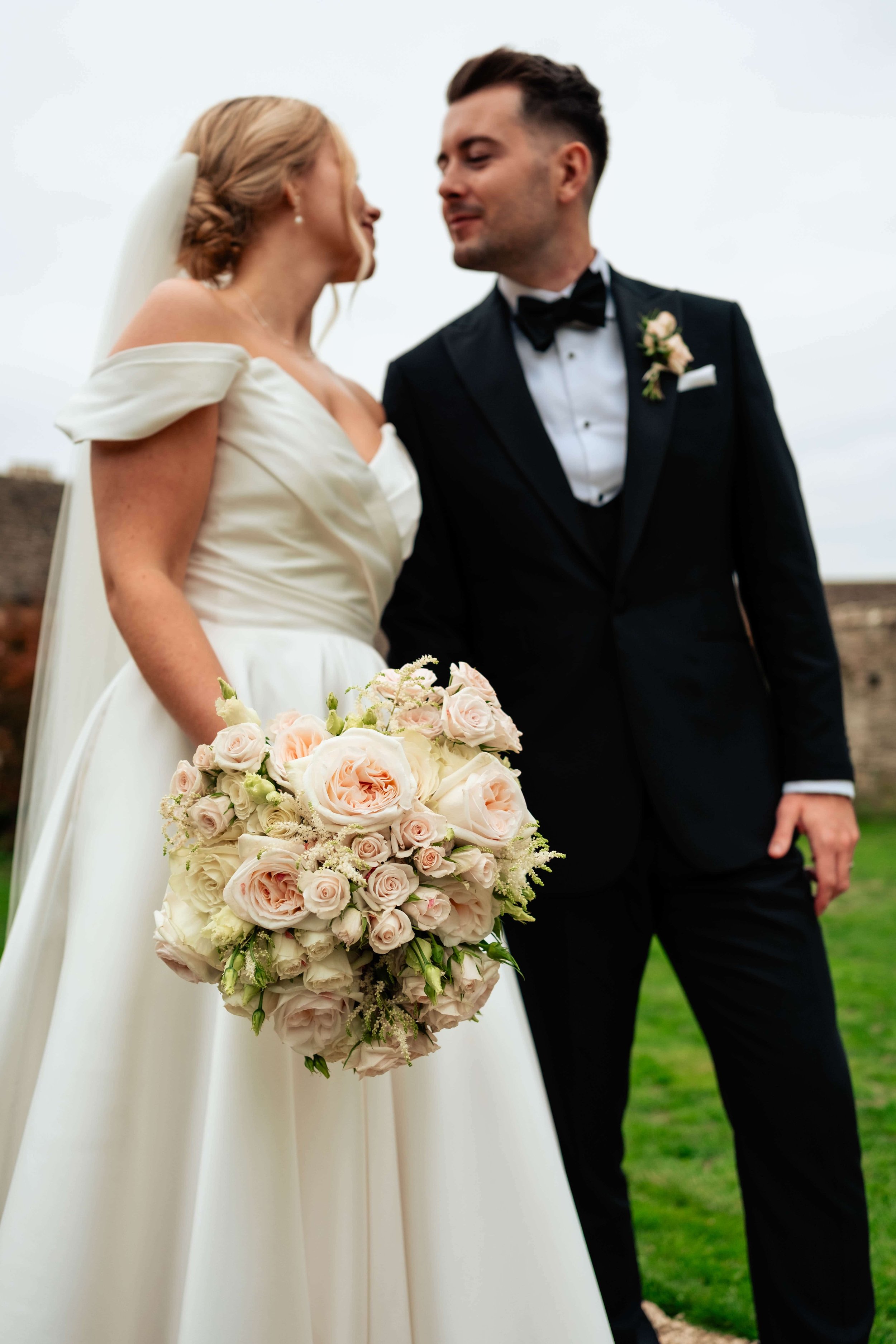 Bride and groom facing each other outdoors, bride holding a bouquet of pink and white roses, bride wearing an off-shoulder white wedding dress, groom in a black tuxedo with bow tie, cloudy sky in background.