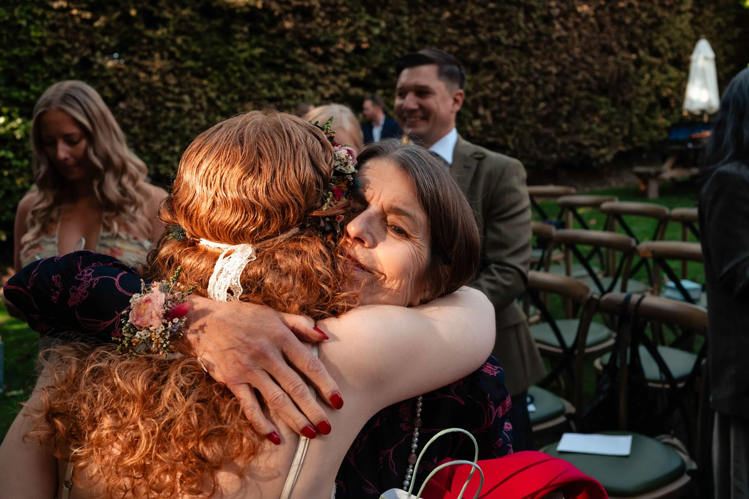 Two women hugging, one with red curly hair wearing a floral headpiece and the other with brown hair in a dark dress, at an outdoor event with seated guests and brown bushes in the background.