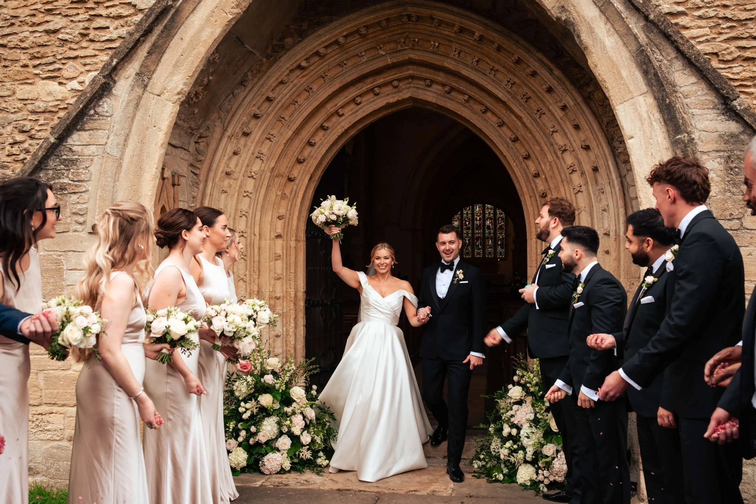 A wedding celebration outside a church, with the bride and groom walking hand in hand through a floral archway, surrounded by bridesmaids and groomsmen.