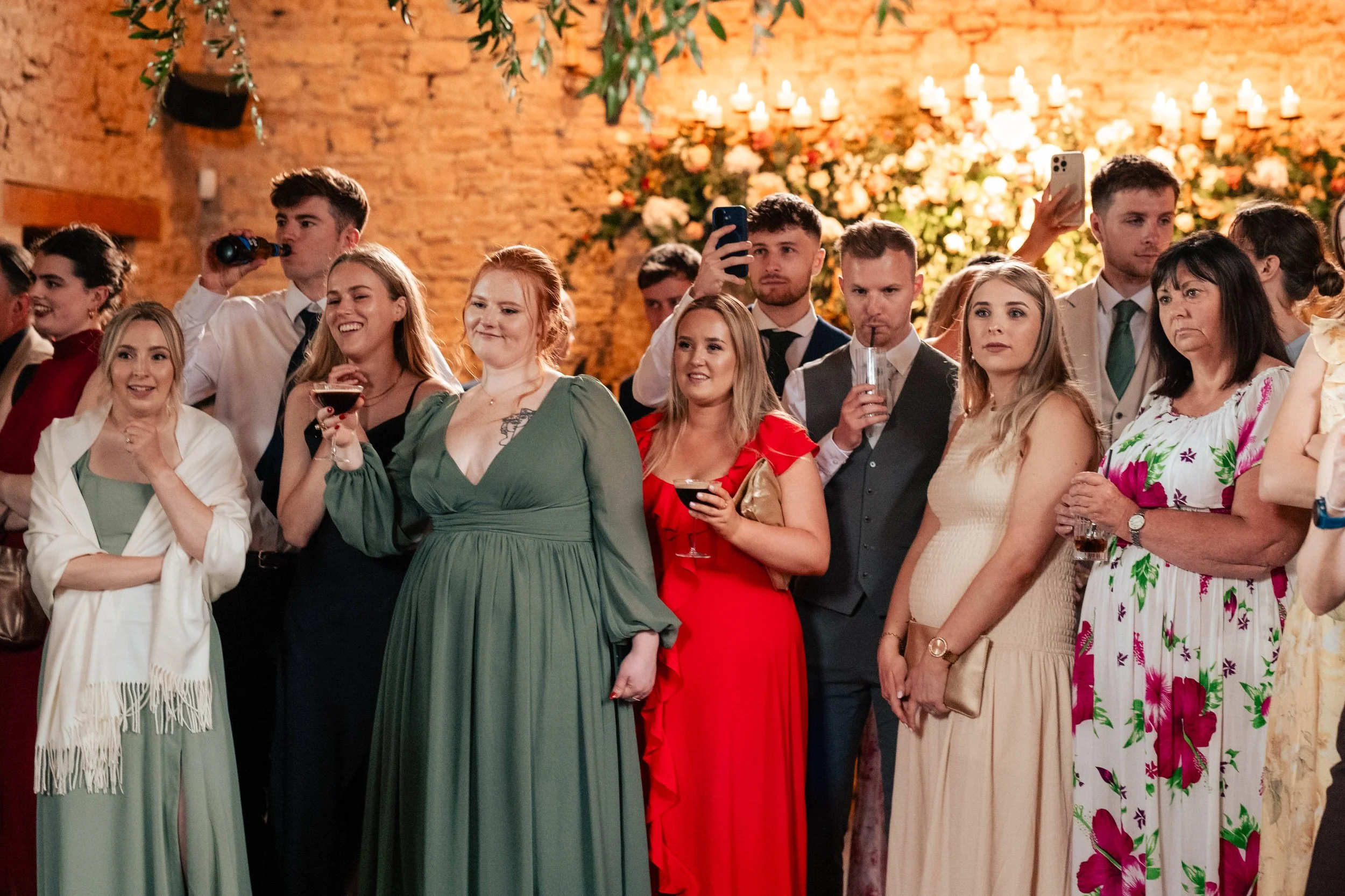 Group of people at a celebration, some holding drinks, in a decorated venue with brick walls and floral arrangements.