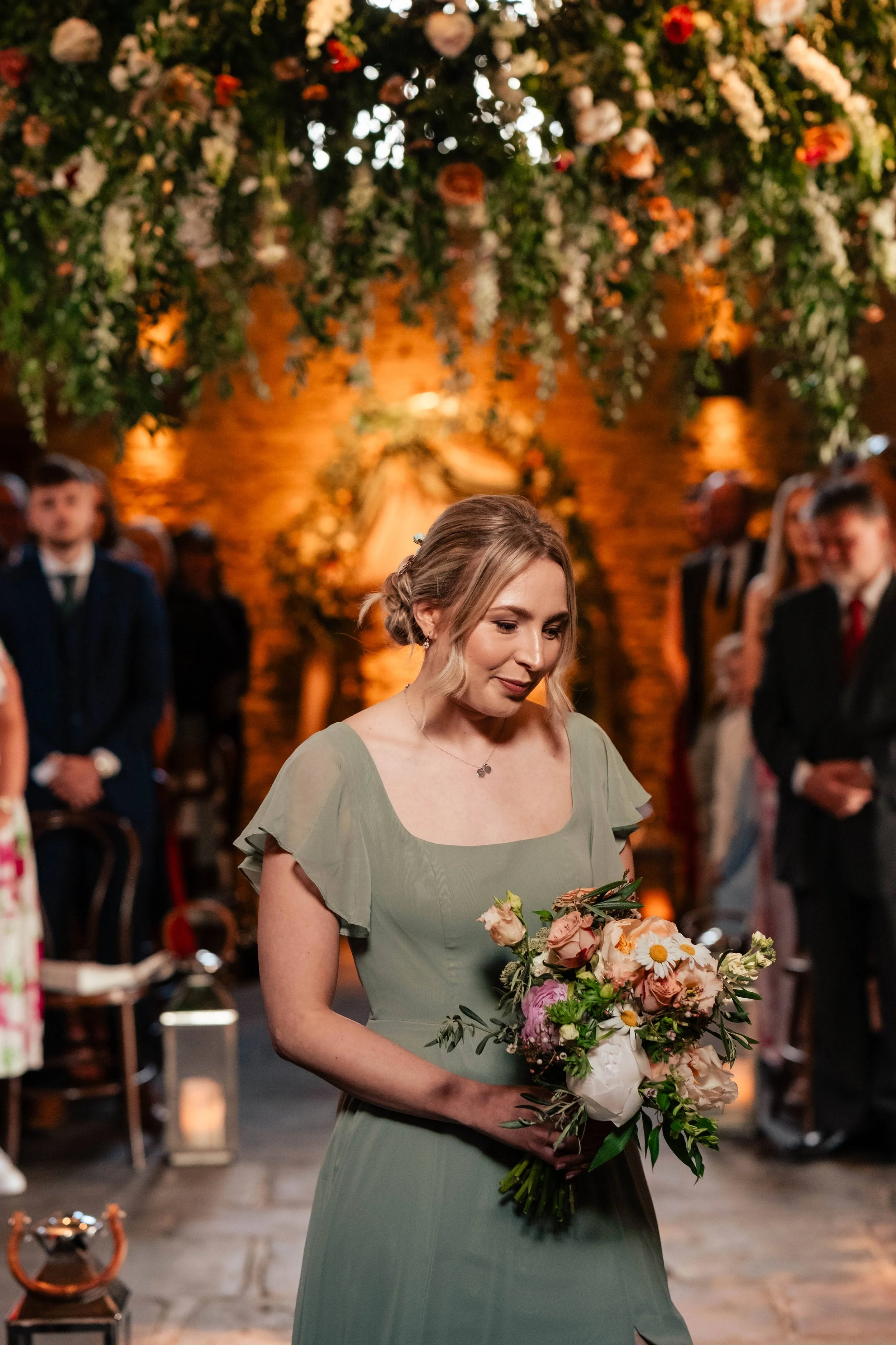 A woman in a green dress holding a bouquet of flowers at a wedding reception with guests in the background and floral decorations hanging above.