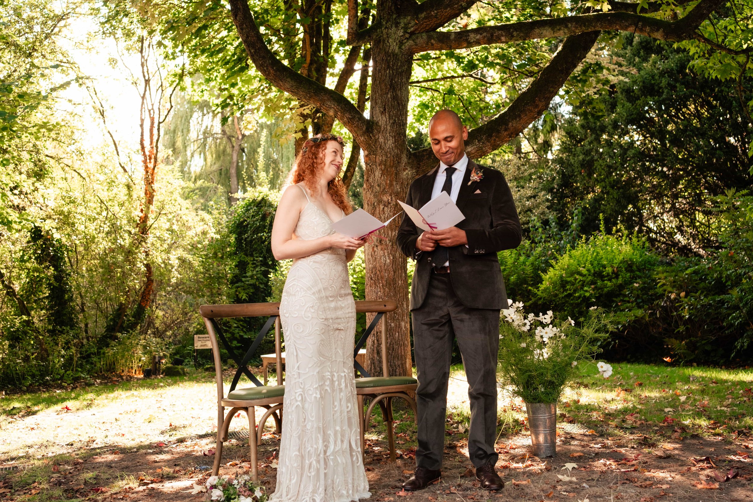 A bride and groom exchanging vows outdoors under a tree, with the bride wearing a white dress and the groom in a black suit. They are holding vows or a script and smiling at each other, with a floral arrangement nearby.