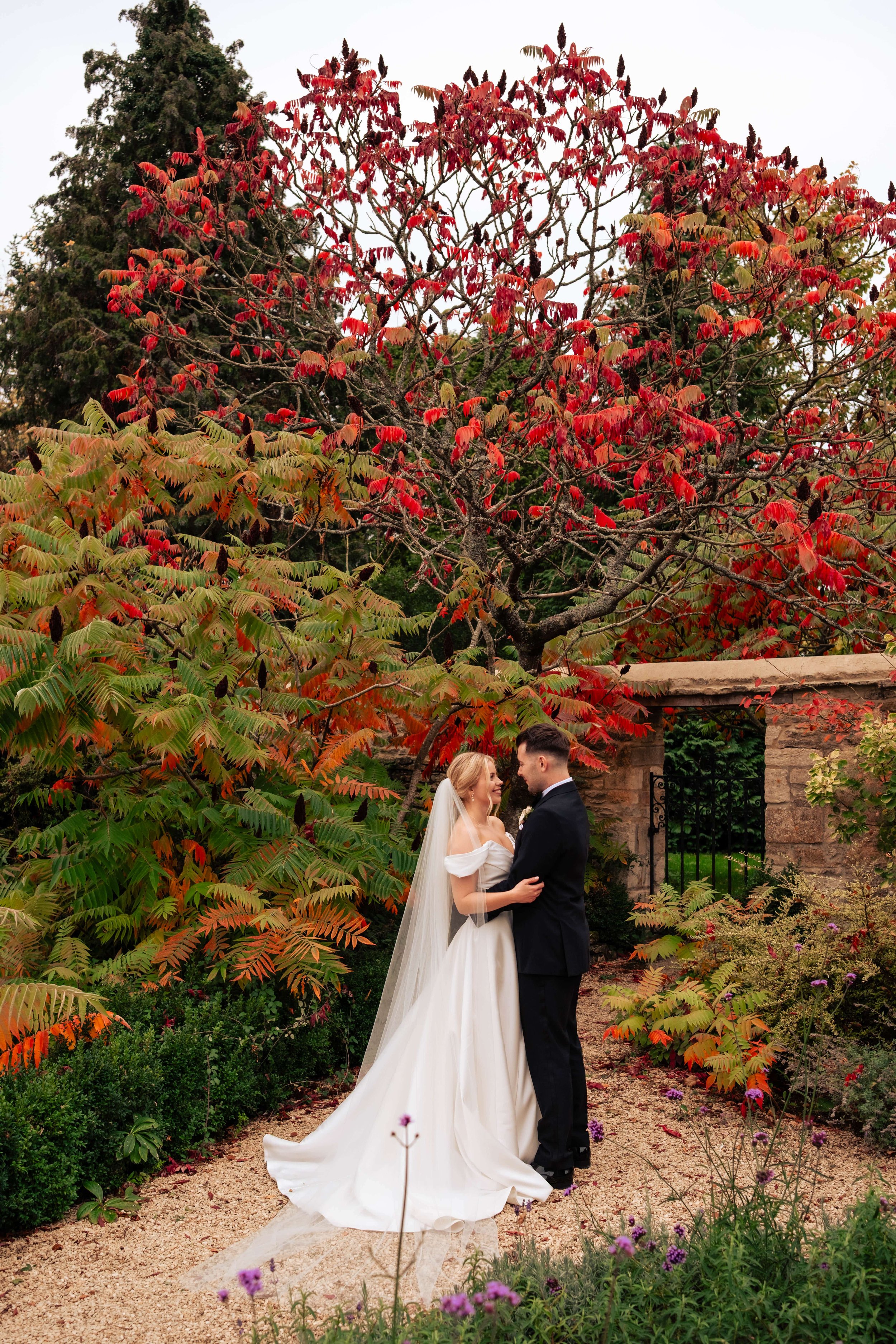 A bride and groom standing close together in a garden with autumn-colored trees and plants, sharing a moment of affection.