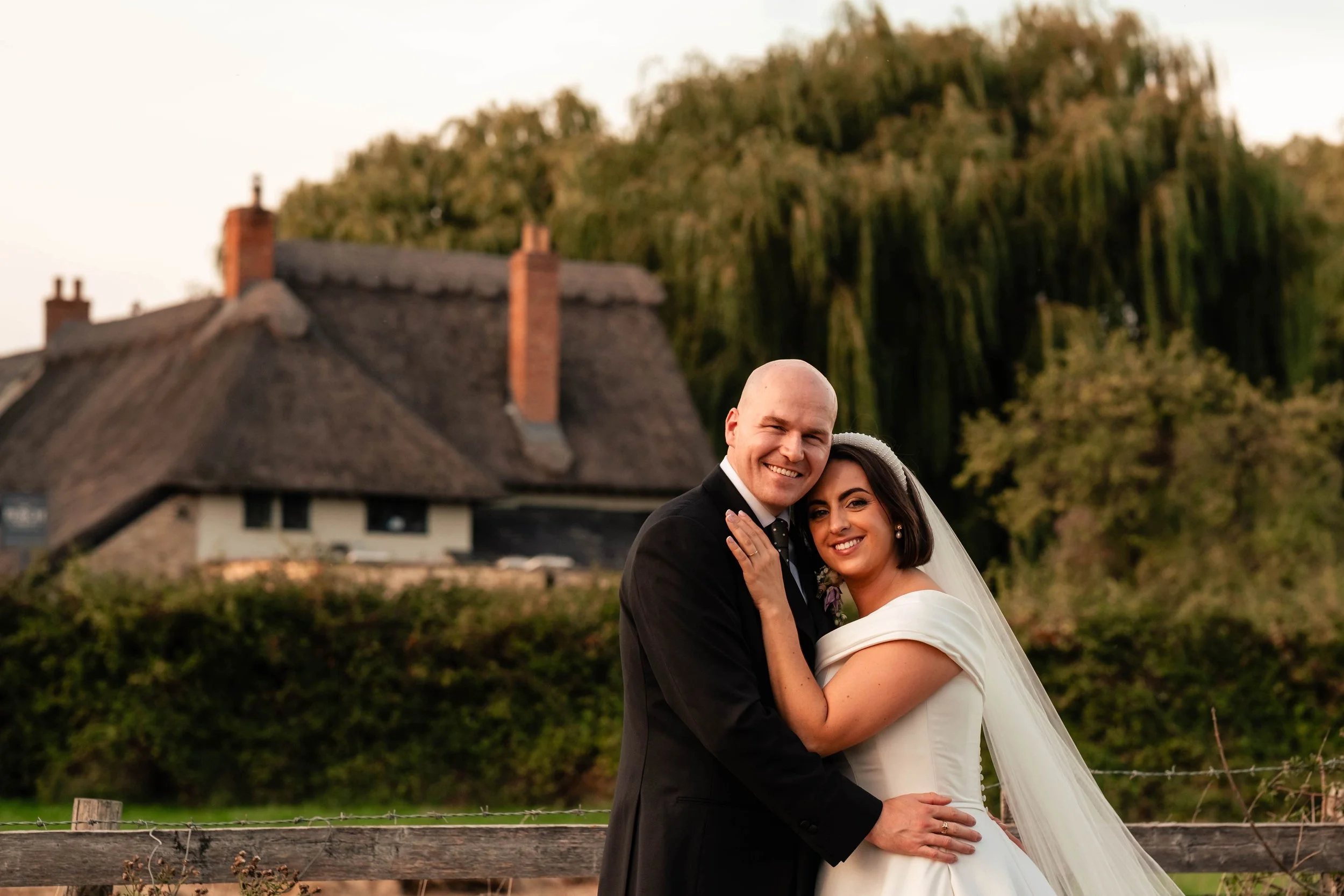 A newly married couple smiling and embracing outdoors near a fence, with a house and trees in the background during sunset.