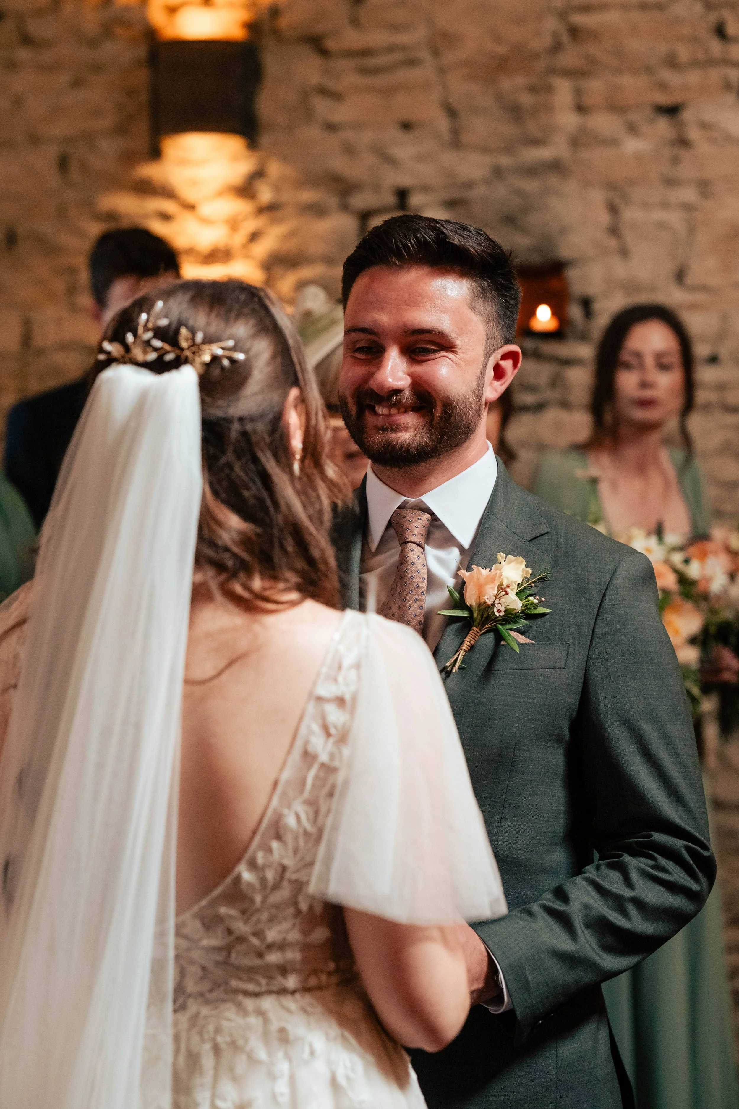 A wedding ceremony with a bride and groom facing each other, smiling, in a rustic indoor setting surrounded by guests.