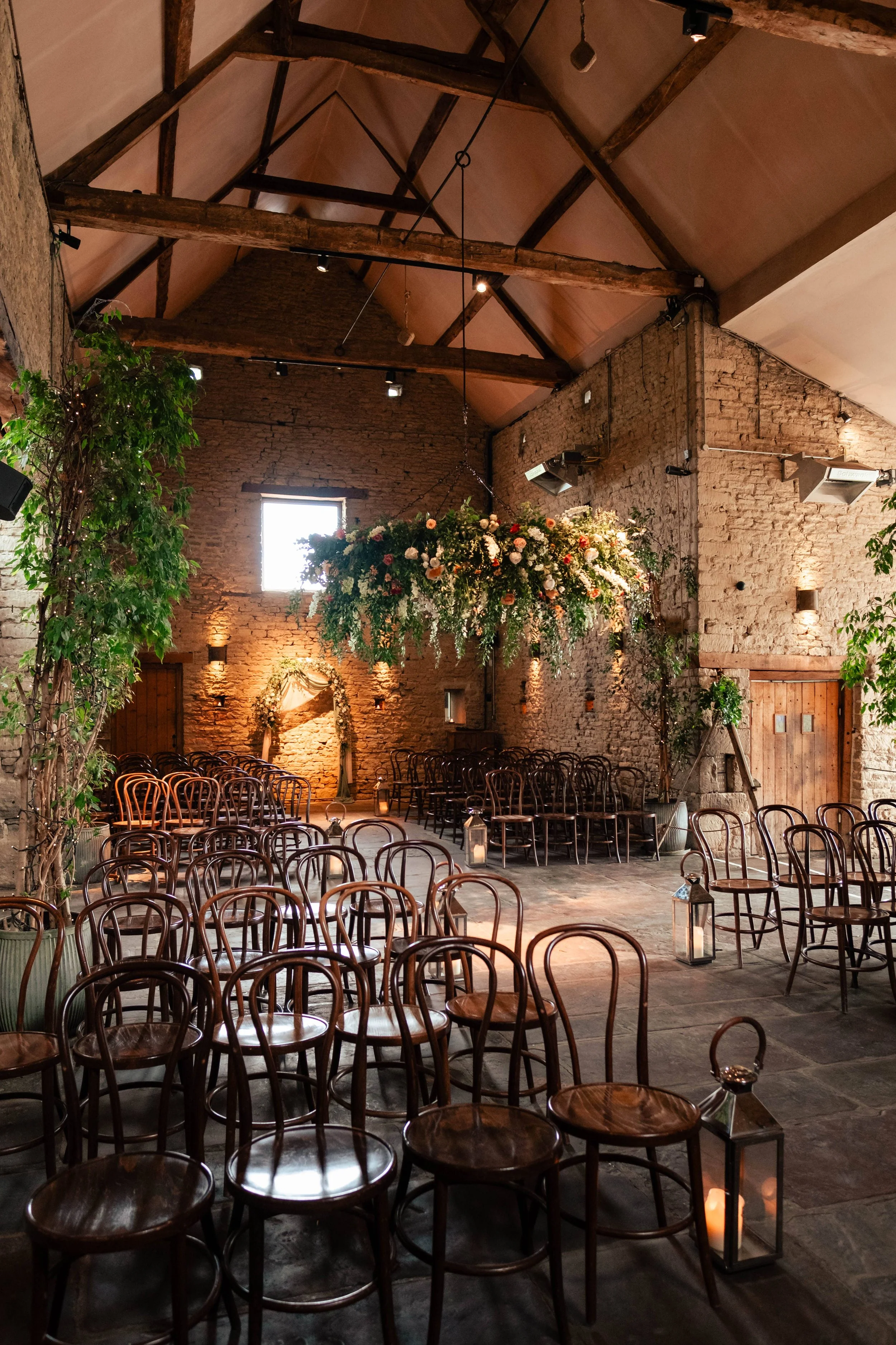 An indoor wedding venue with wooden chairs arranged for a ceremony, decorated with lanterns, large floral chandelier, rustic brick walls, and exposed wooden beams in the ceiling.