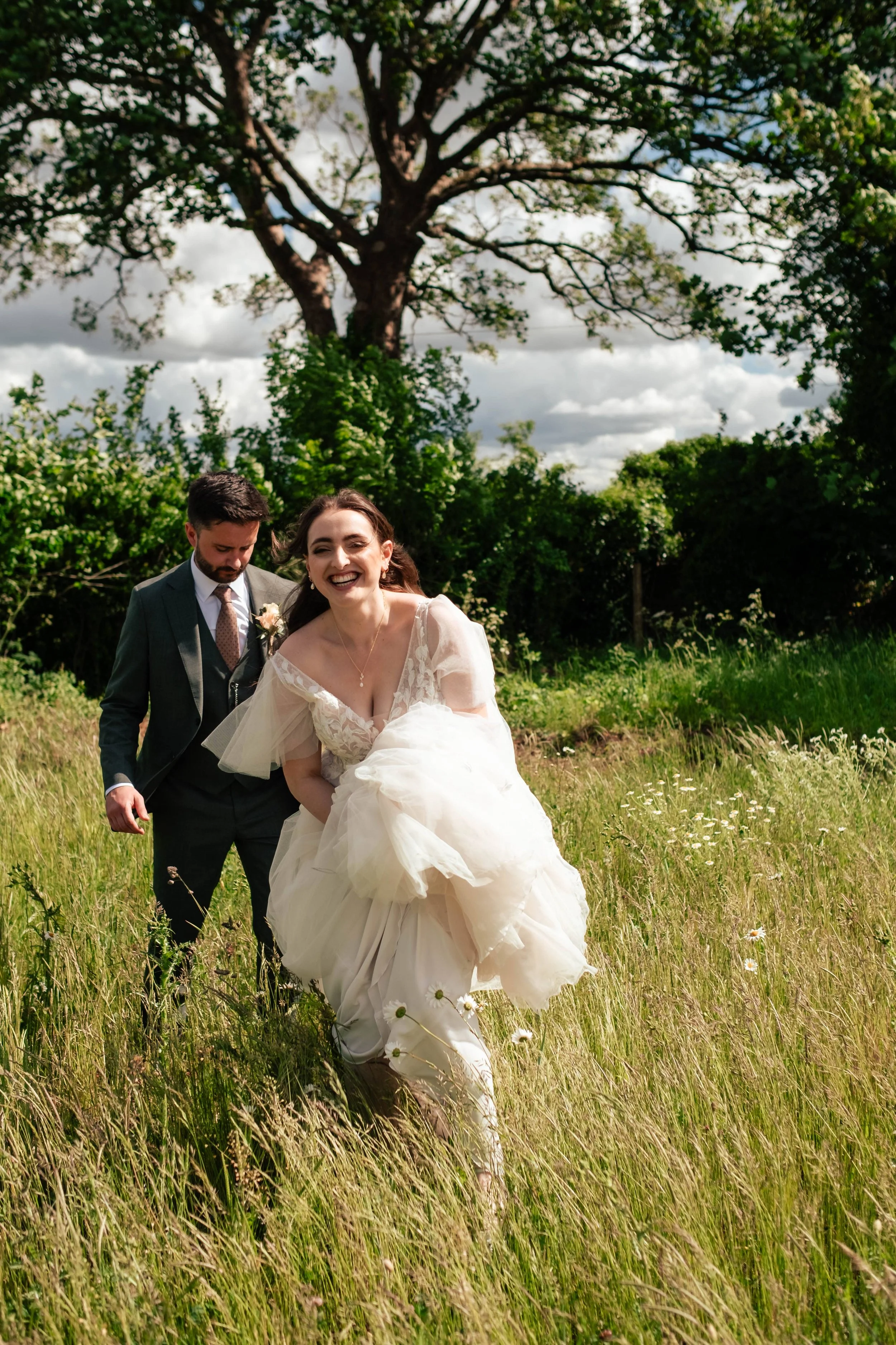 A bride and groom running through a grassy field on their wedding day, smiling and joyful.