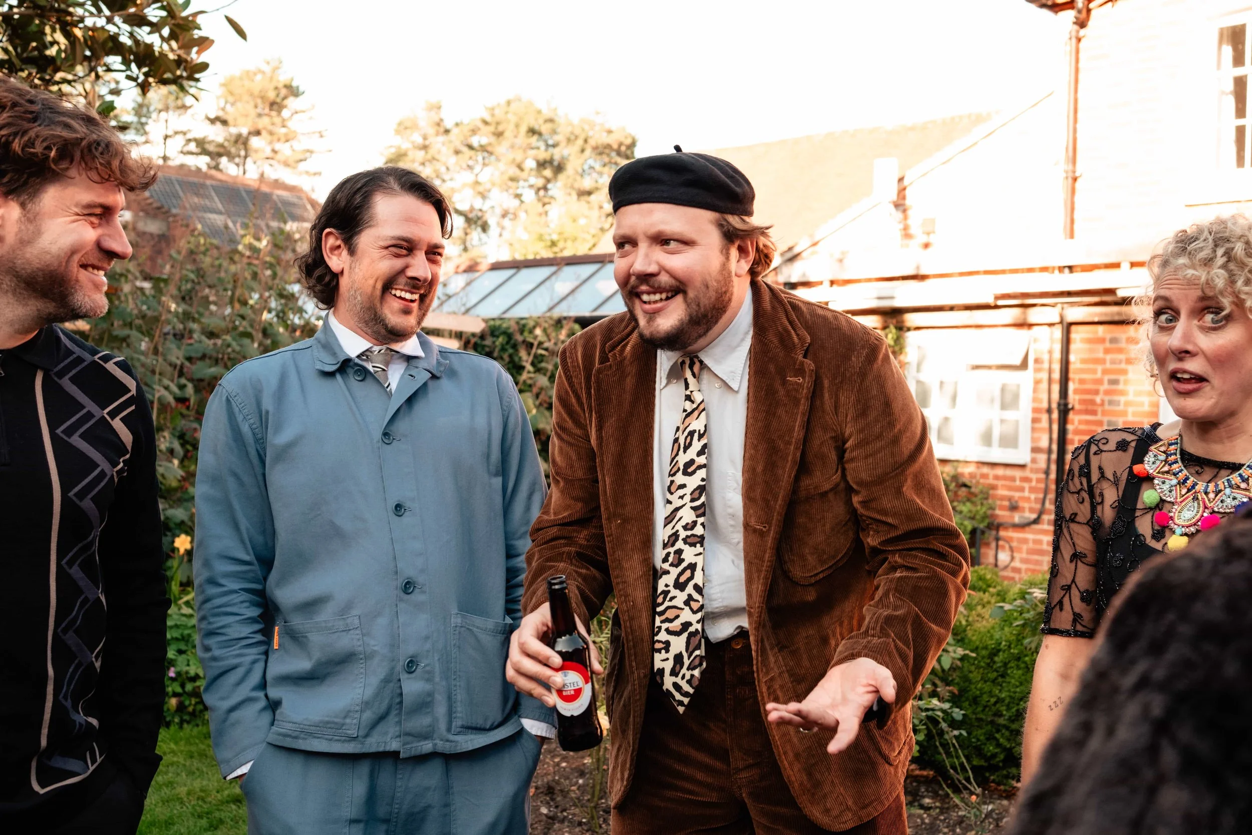 Group of five people smiling and talking outdoors in a garden during daytime, with a brick house and greenery in the background.