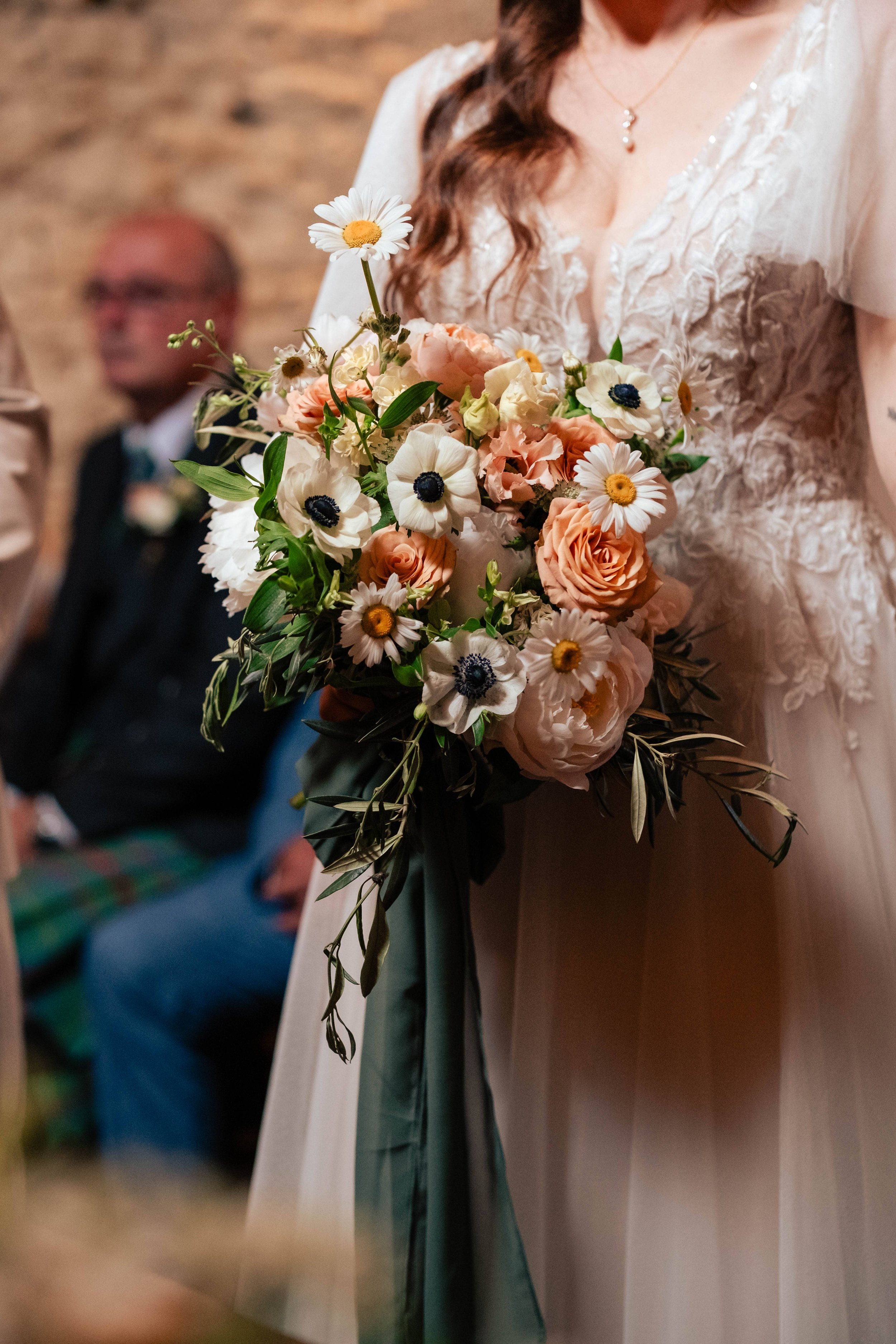A woman in a lace dress holding a bouquet of white, peach, and cream flowers with a dark green ribbon at a wedding ceremony.