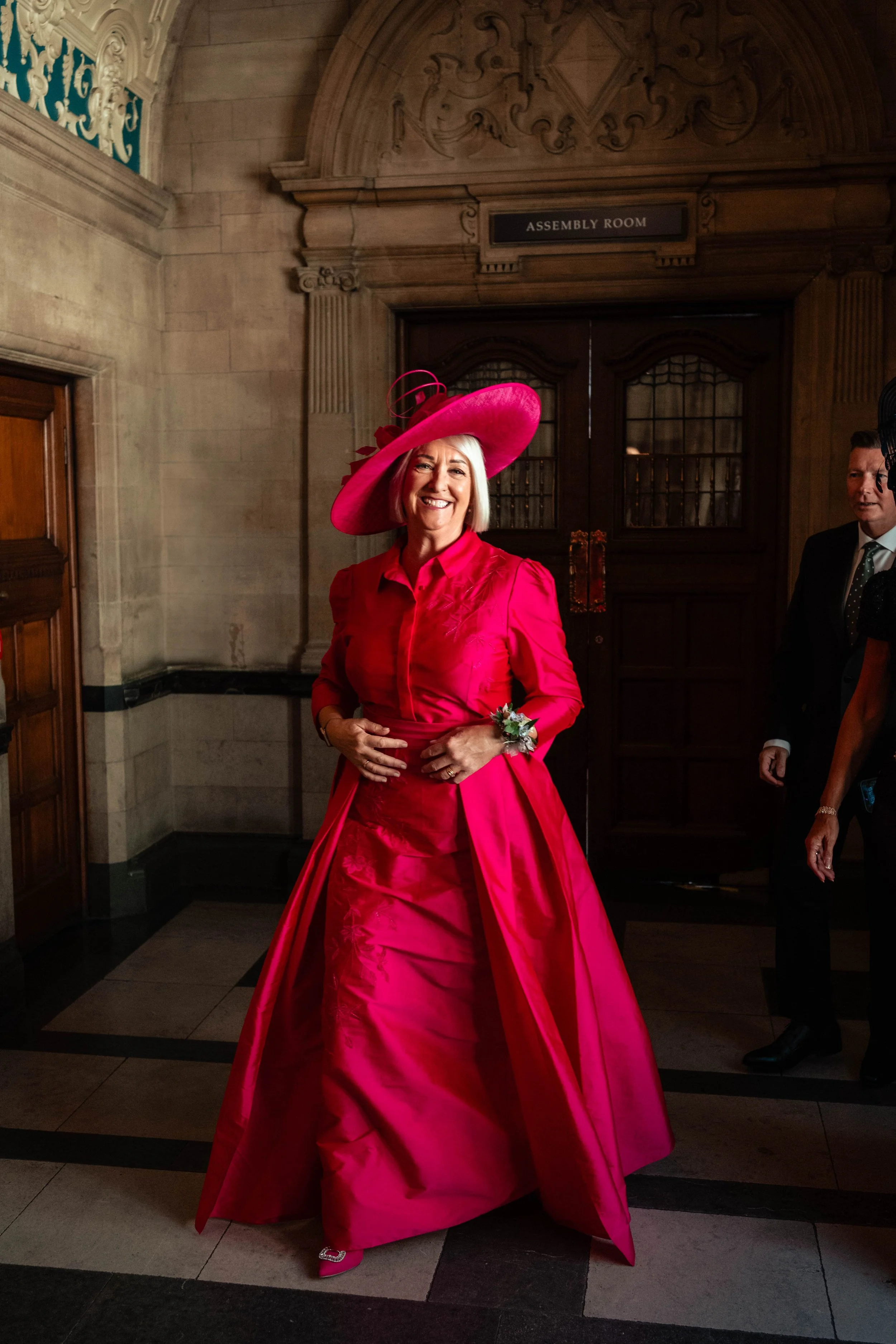 A woman in a bright pink dress and large pink hat smiling indoors in front of a wooden double door marked "Assembly Room."