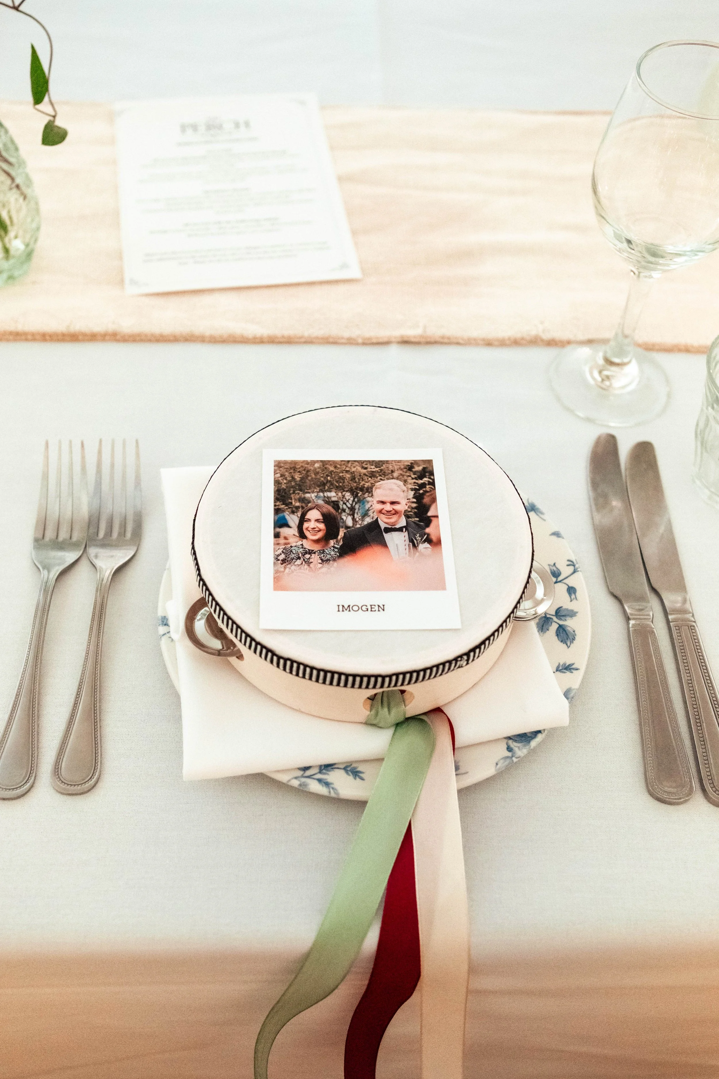 A wedding table setting with a plate, white napkin, and ribbons in green, cream, and red. A framed photo with two smiling people and the word 'IMOGEN' underneath is placed on top of the plate. Silverware is arranged on both sides of the plate, and em