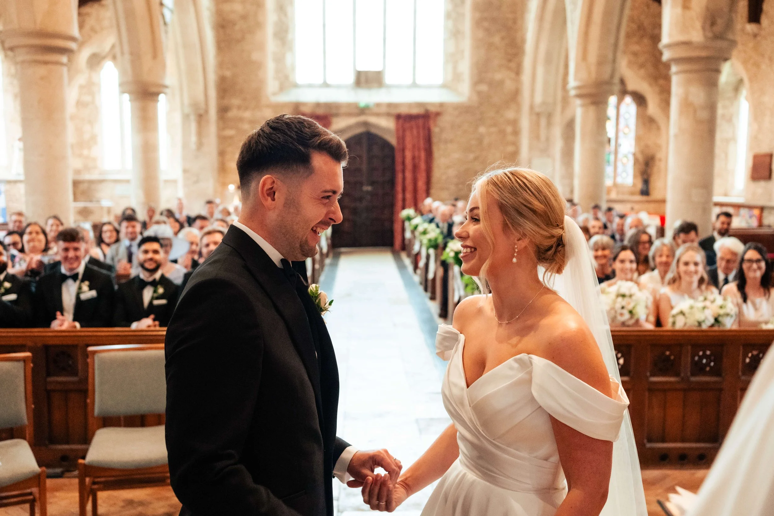 A bride and groom holding hands and smiling during their wedding ceremony in a church filled with seated guests.