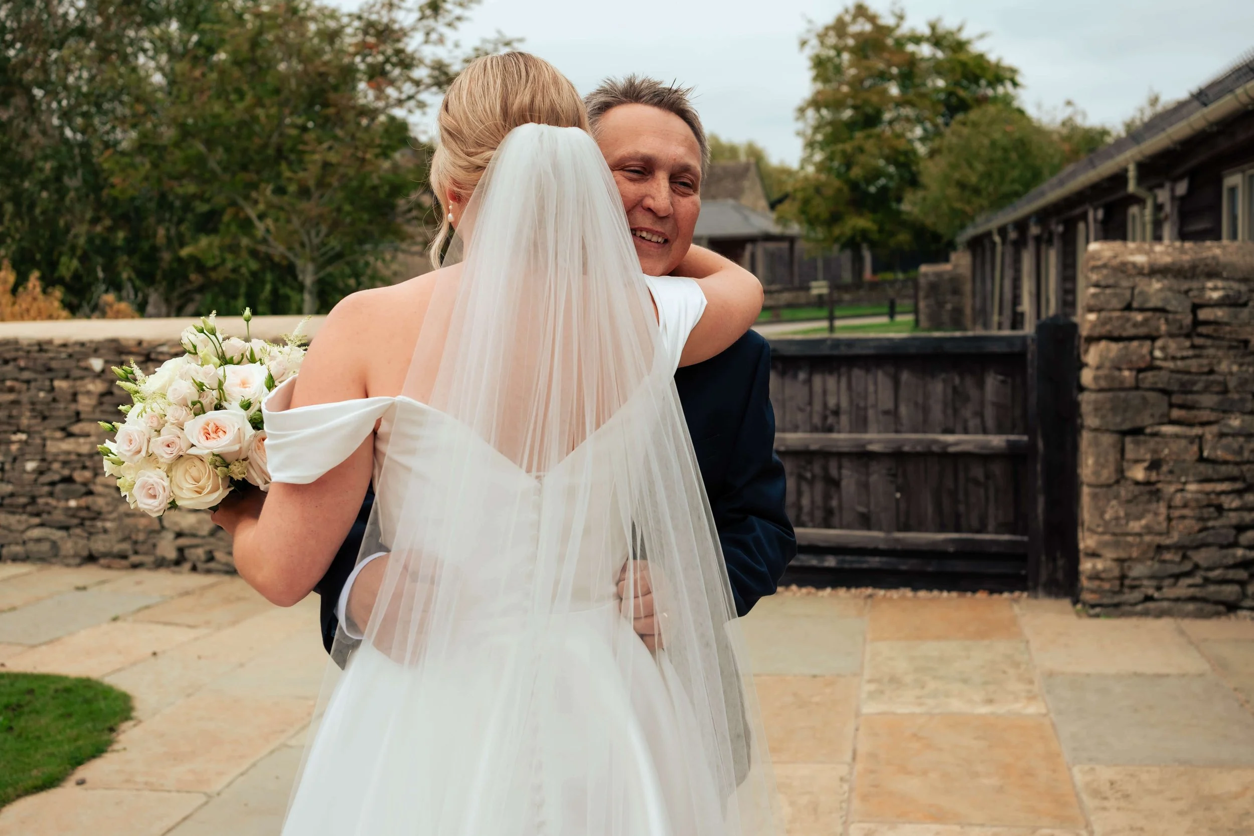 A bride and her father, sharing a hug outdoors on her wedding day. The bride is holding a bouquet of white and blush roses, wearing a white off-shoulder wedding gown and a veil. The background features a stone wall, trees, and historic buildings.