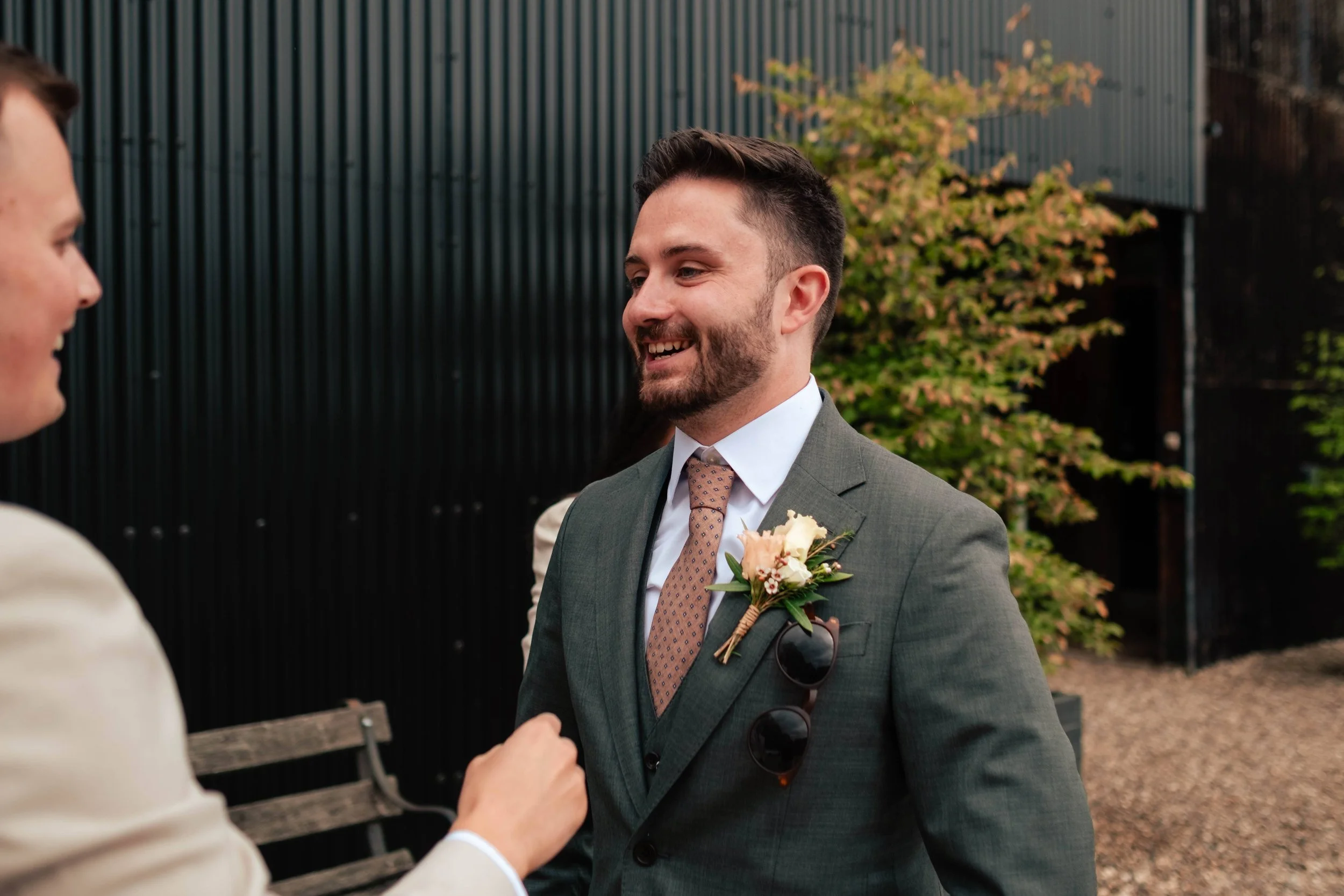 A man with a beard in a green suit wearing a boutonniere, smiling and talking to a woman outside during a formal event.