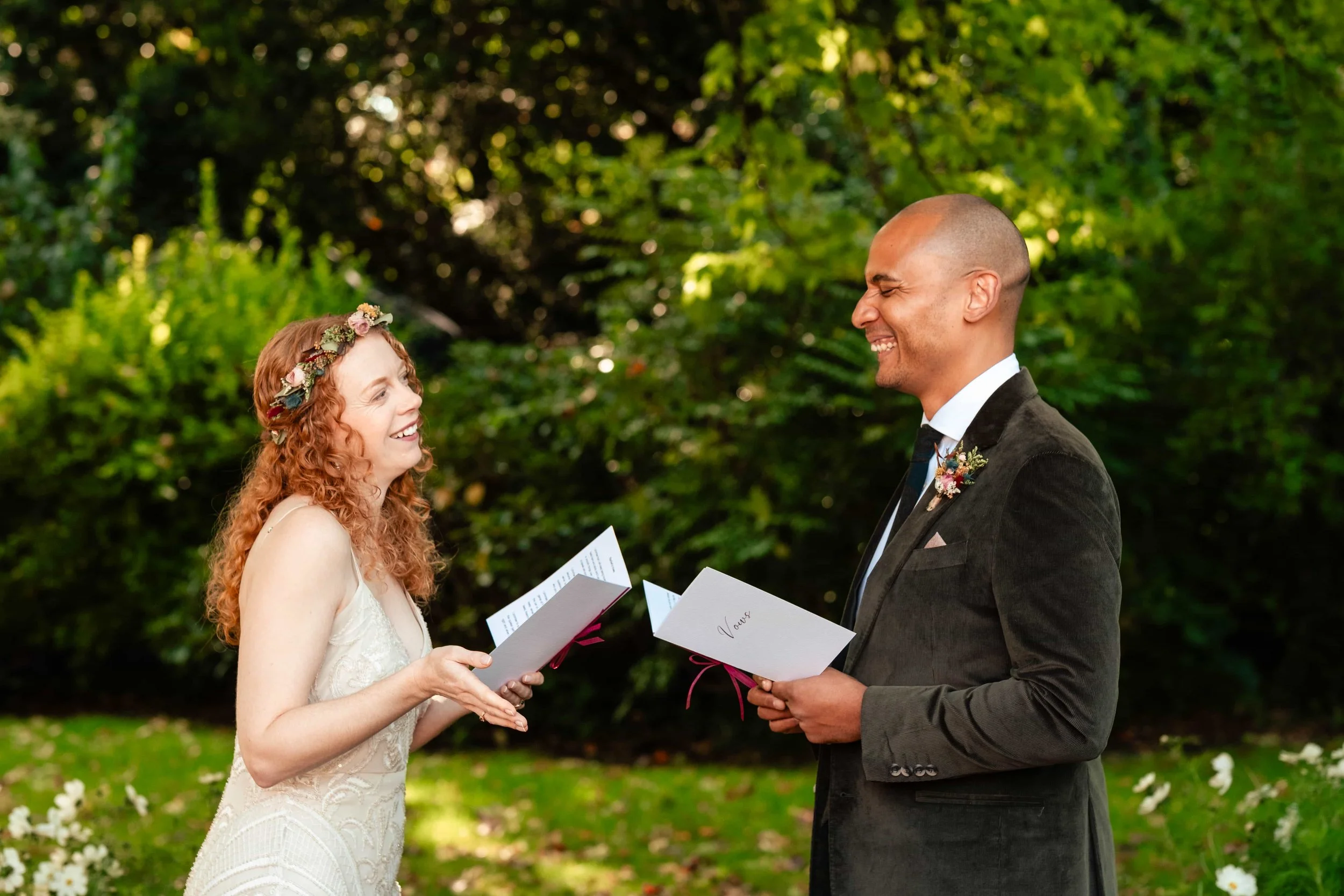 A bride and groom exchanging vows outdoors, with the bride smiling and the groom laughing, surrounded by greenery and flowers.