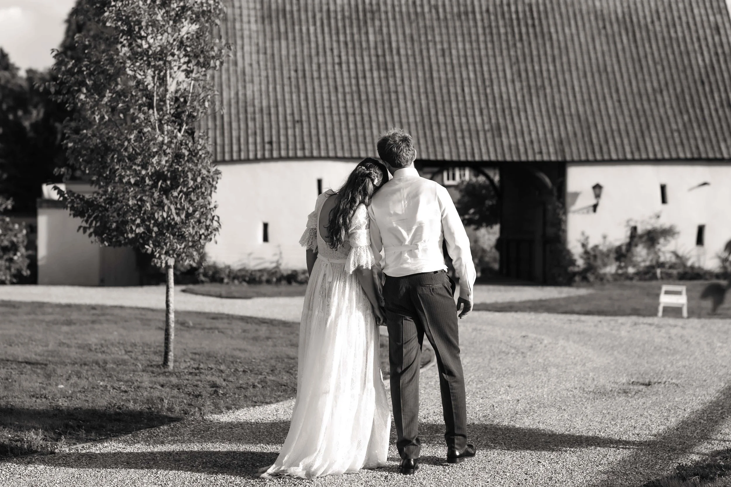 A black and white photo of a bride and groom walking together outside near a building with a sloped roof, holding hands. The bride is resting her head on the groom's shoulder.