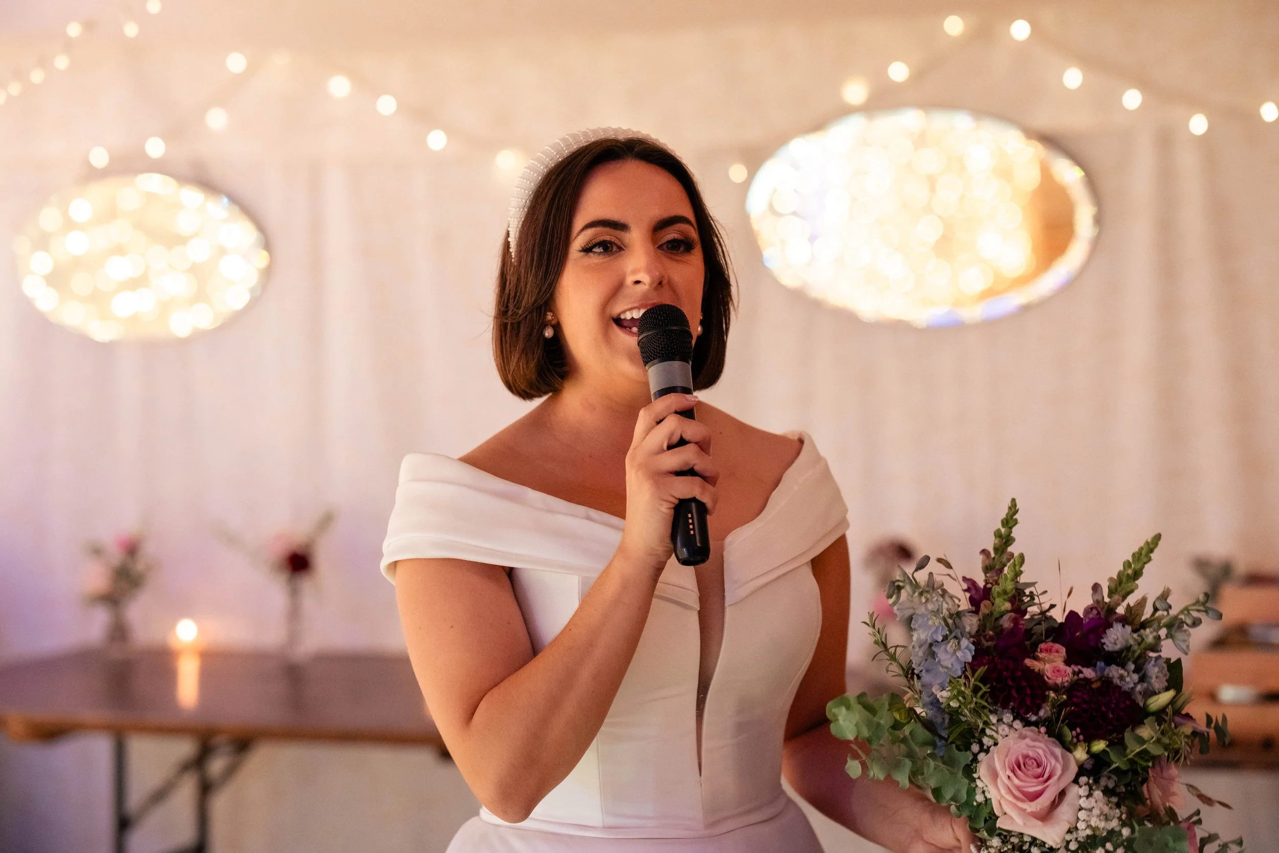 A woman in a white wedding dress holding a bouquet of flowers and speaking into a microphone at her wedding reception.