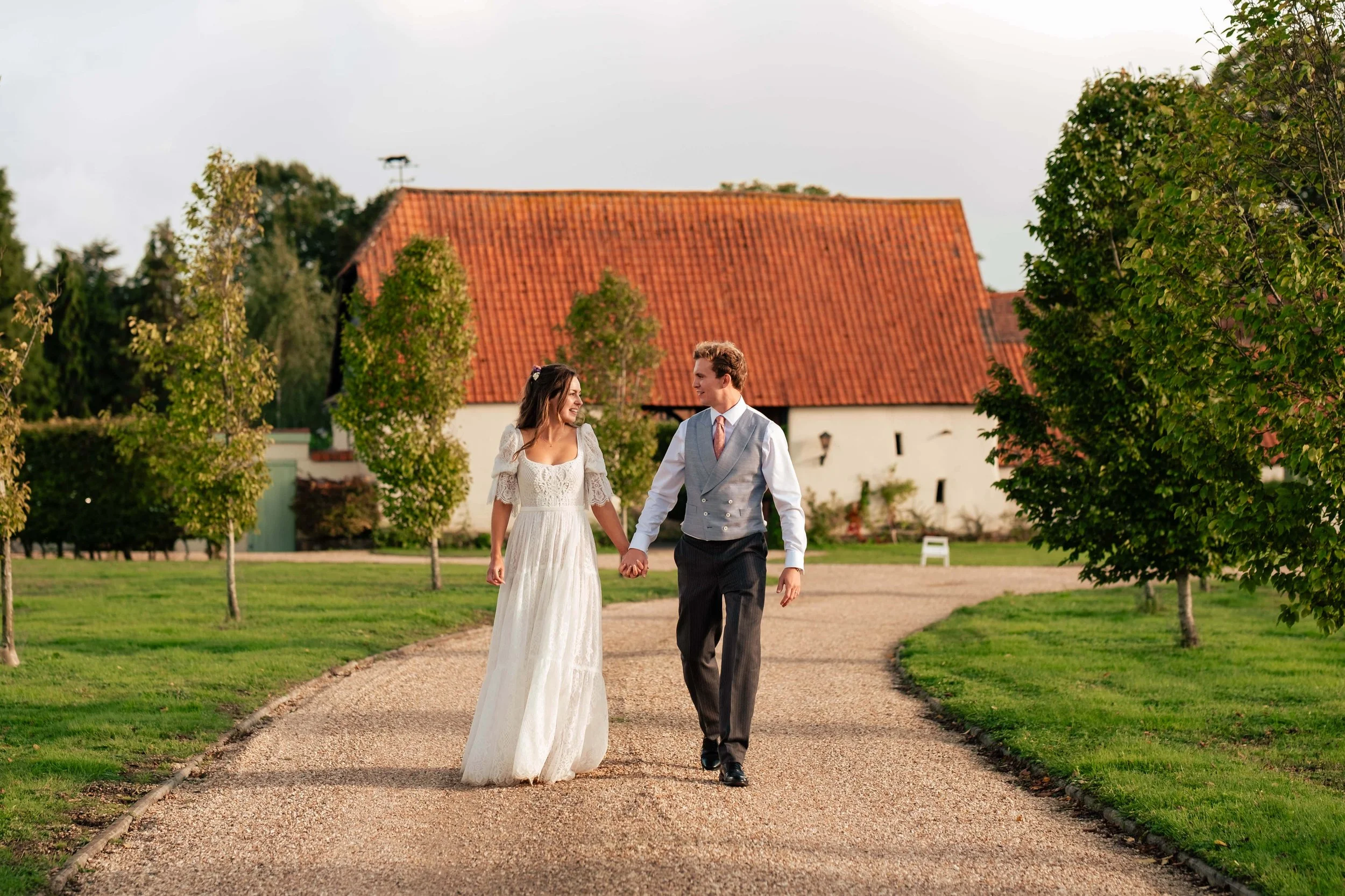 A couple walking hand in hand on a gravel path in a park-like setting, with a white house and trees in the background, during daytime.