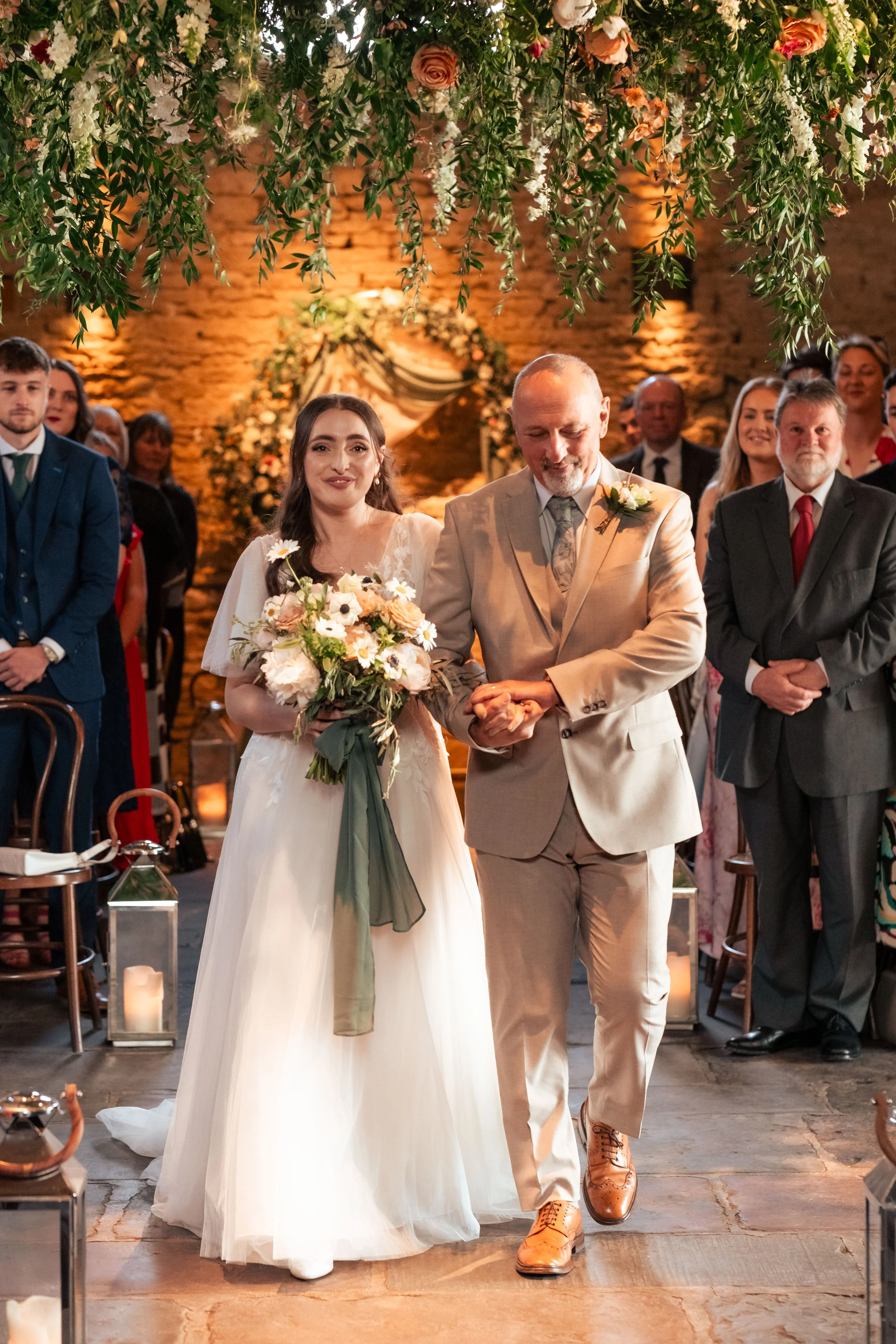 A bride in a white wedding dress holding a bouquet of flowers walking down the aisle with an older man in a beige suit at a wedding ceremony.