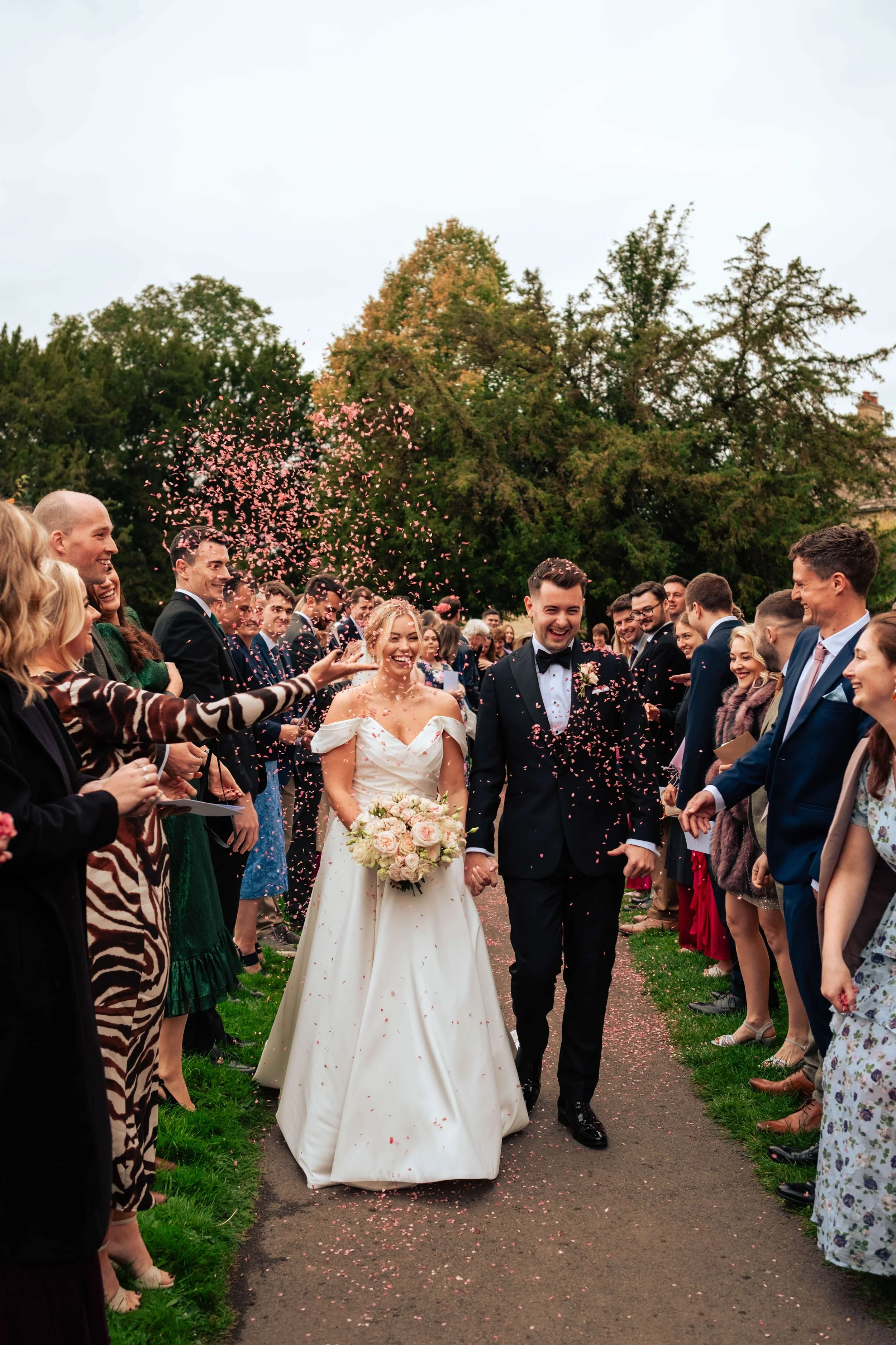 A bride and groom walking down a path, surrounded by guests celebrating their wedding with confetti and smiles, outdoors on a cloudy day.