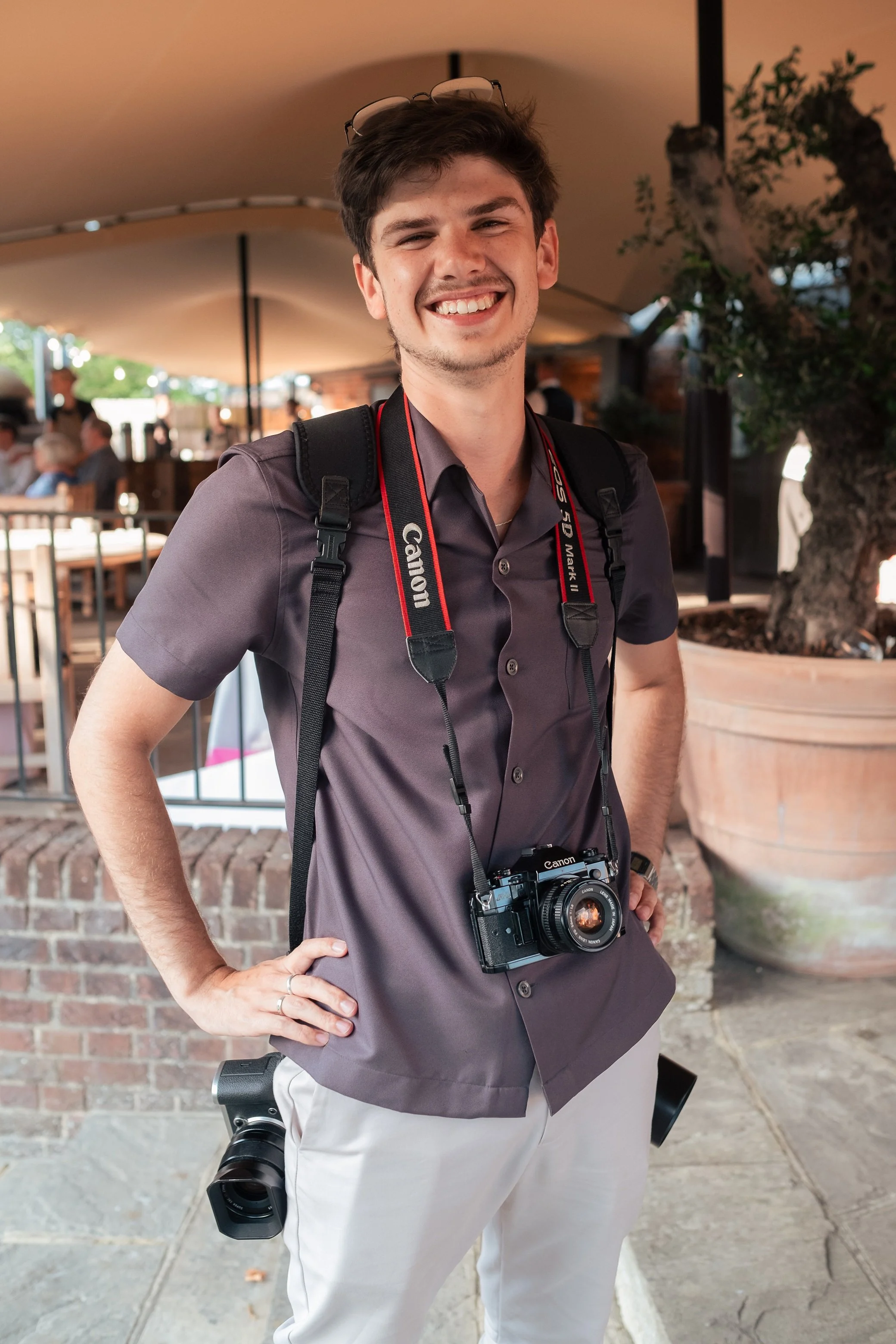 A young male photographer with dark hair and a wide smile standing outside, wearing a gray shirt, cream trousers, and carrying multiple cameras and a backpack.