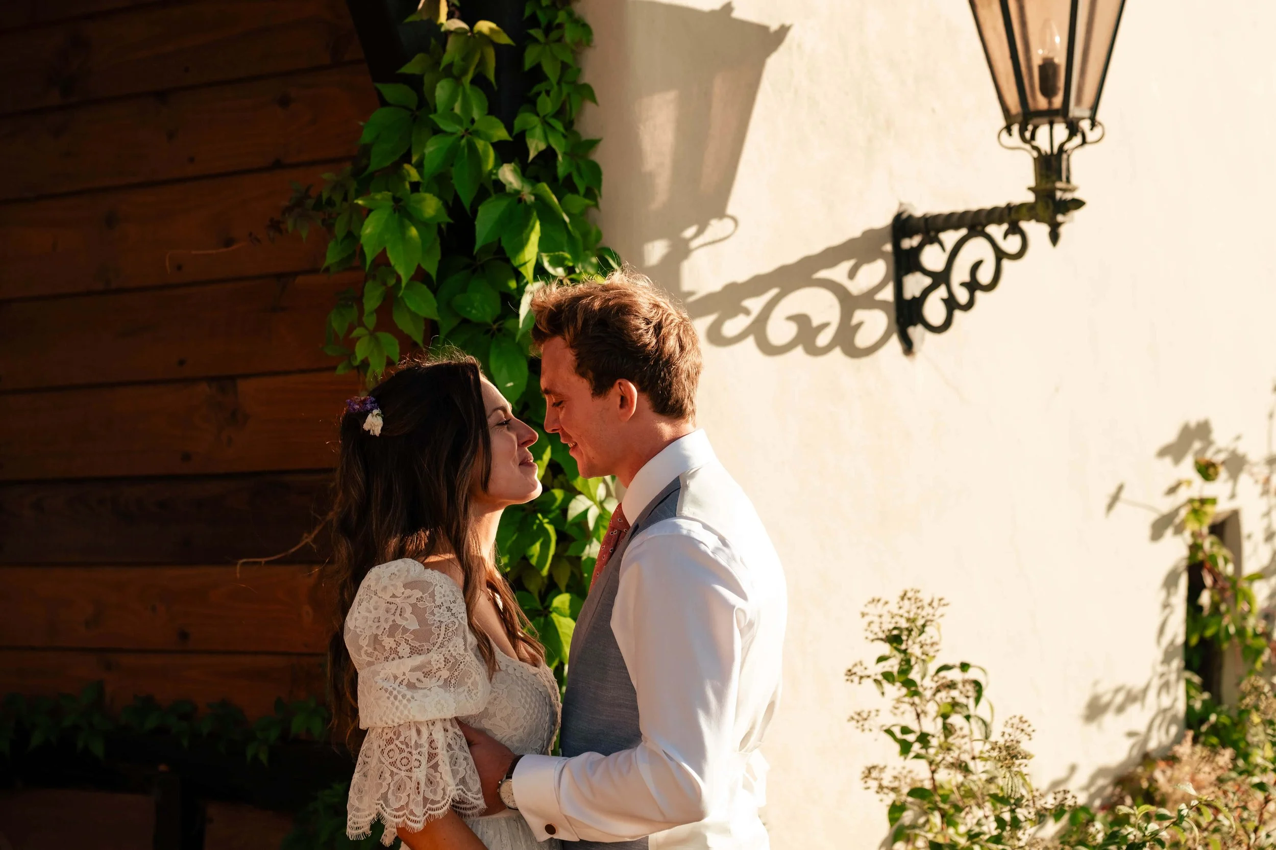 A bride and groom standing close together outdoors, facing each other with foreheads touching, smiling in a romantic moment during sunset, with a white wall and green plants in the background.