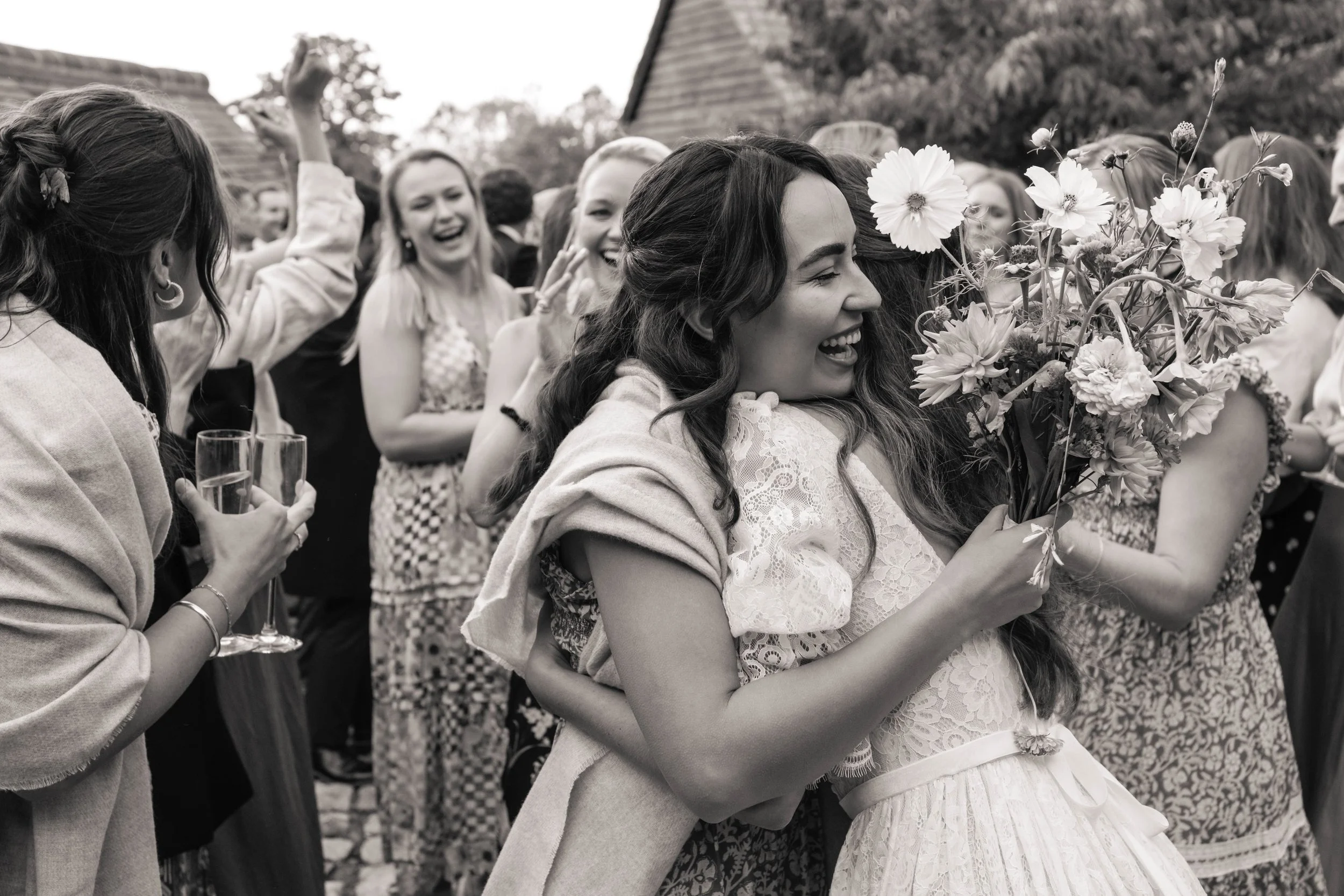 A group of women celebrating outdoors, with a bride in a white dress holding a bouquet of flowers, surrounded by smiling friends who are cheering and clapping.