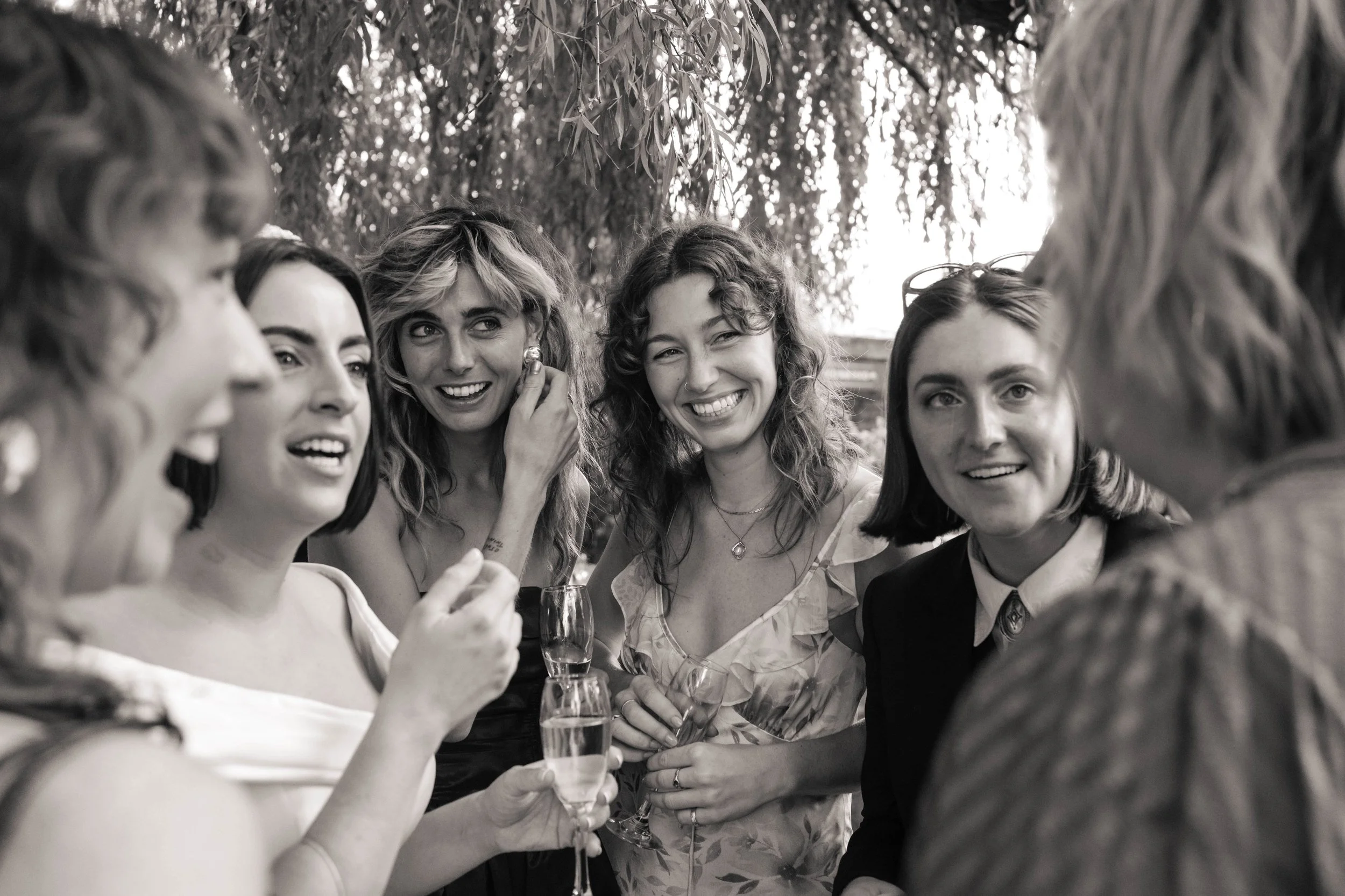 Six women gathered outdoors, smiling and talking at a social event, some holding glasses of champagne.