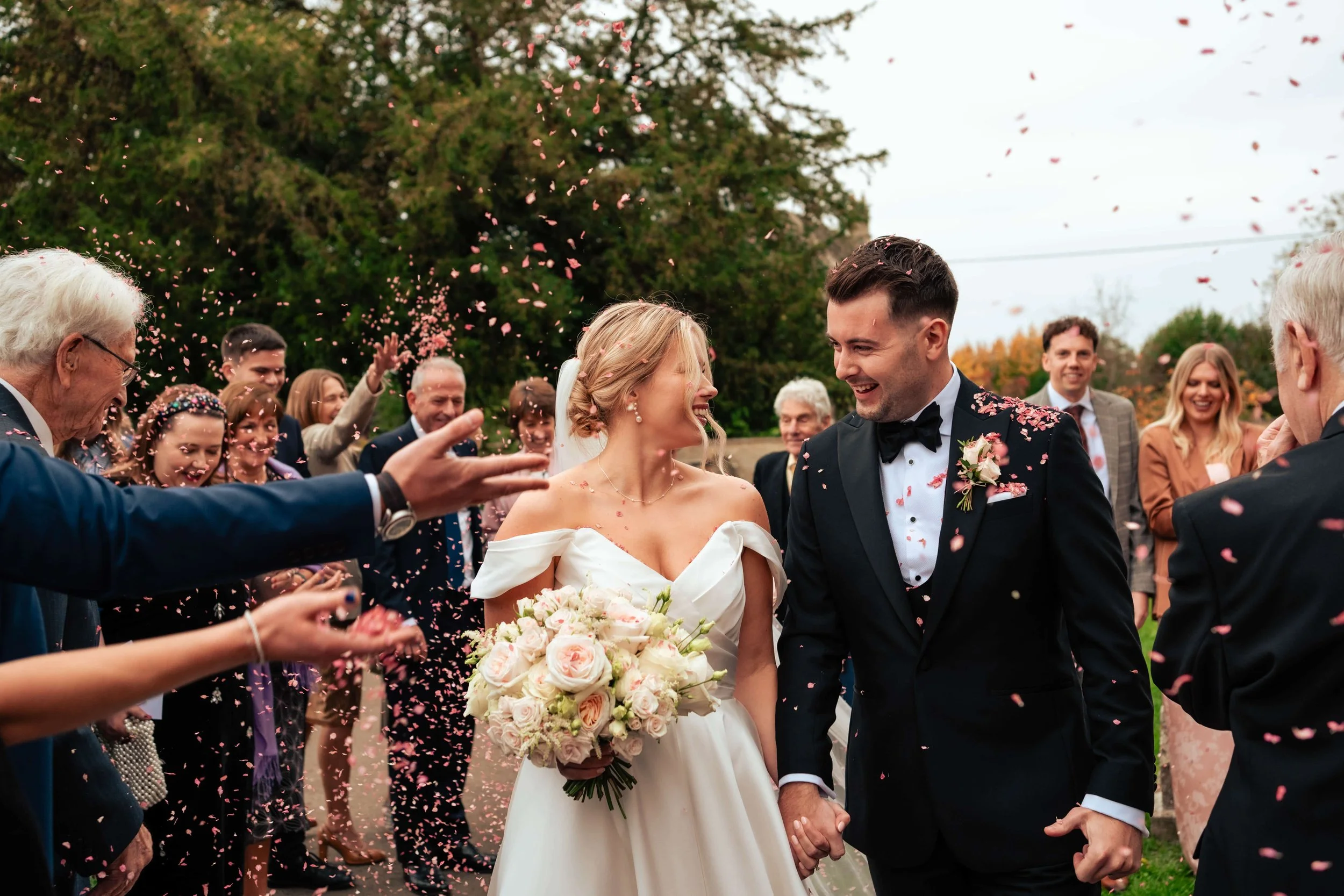 A newlywed couple holding hands and smiling at each other during their outdoor wedding celebration, with guests throwing pink confetti behind them.