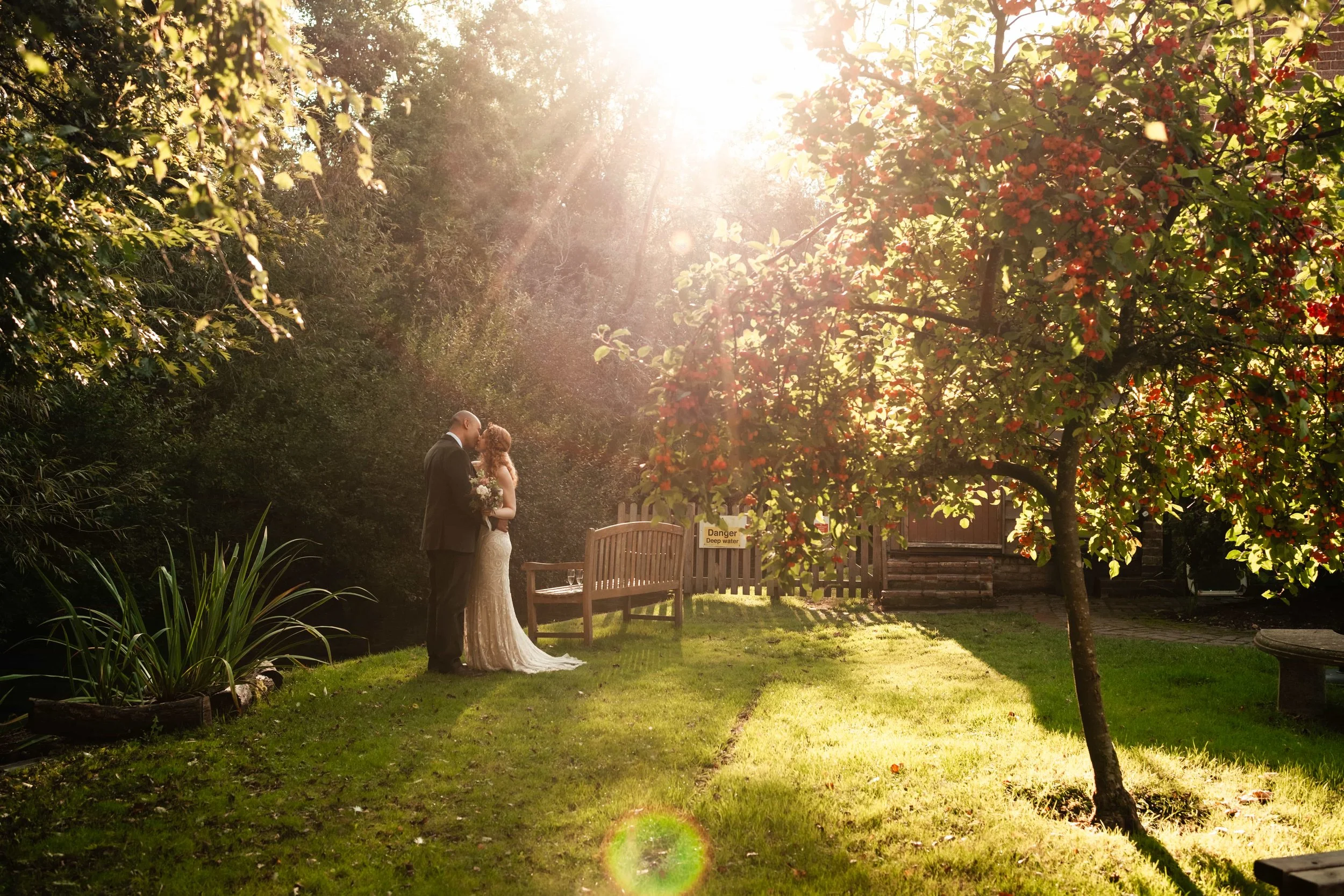 A bride and groom standing close together in a garden, kissing, with sunlight shining overhead and trees, including one with red leaves, around them.