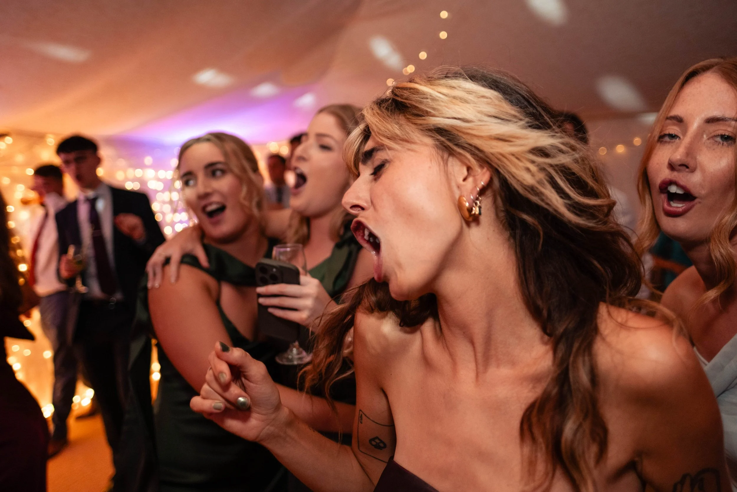 Group of women singing and dancing at a party or celebration, with colorful lights and friends in the background.