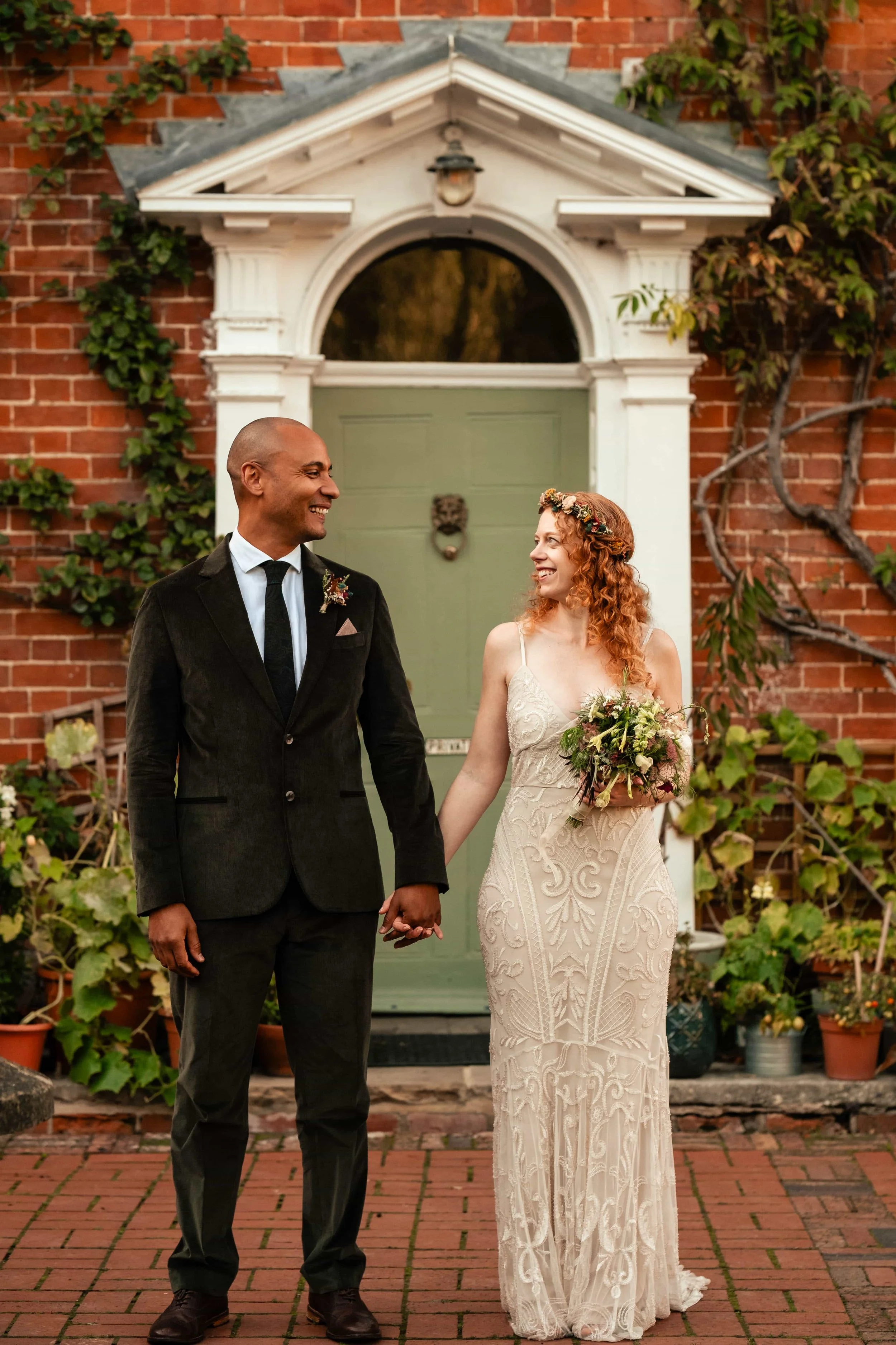 A newlywed couple holding hands and smiling at each other in front of a green door with a red brick wall and plants around them.