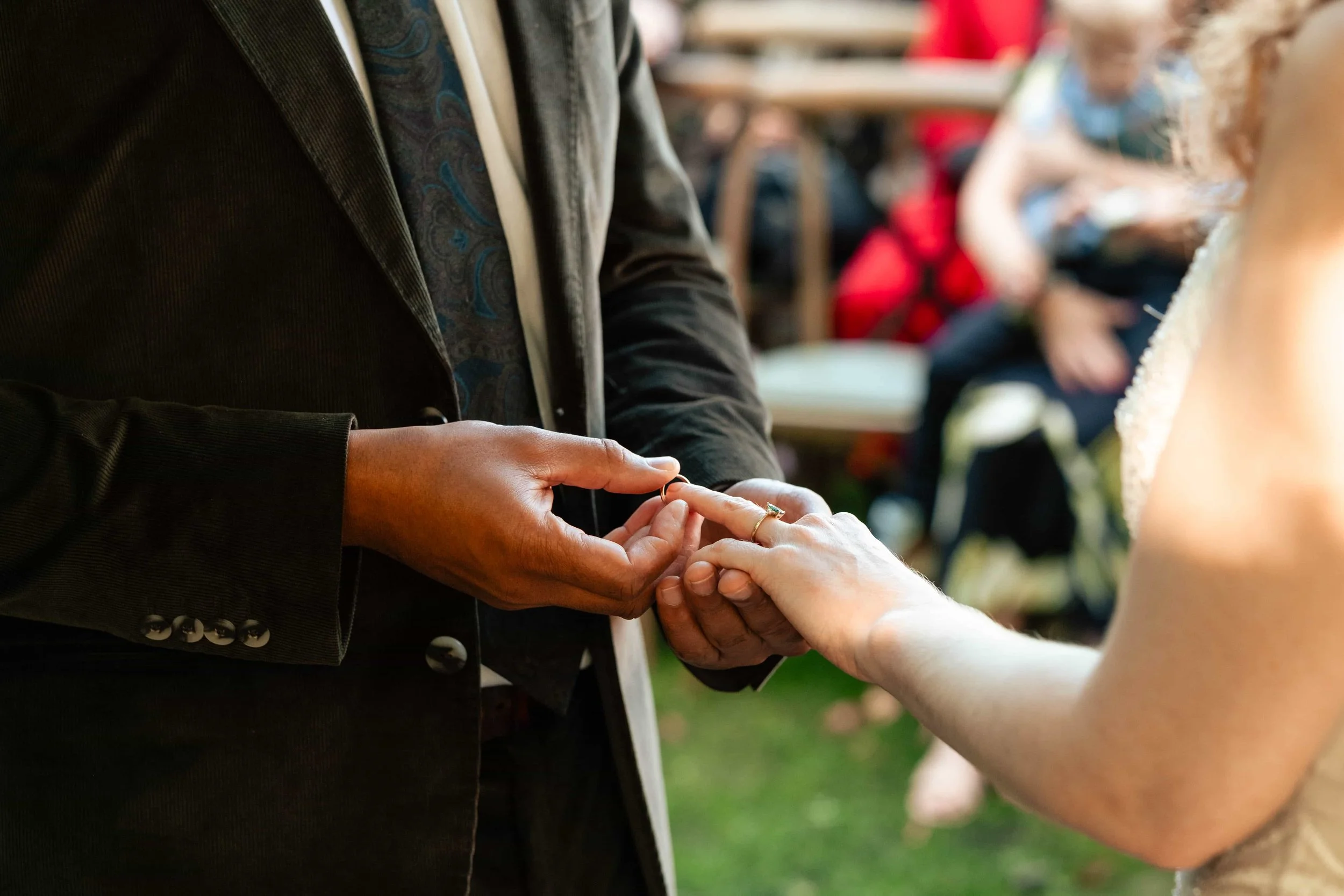 Person exchanging wedding rings during a wedding ceremony outdoors, with guests in the background.